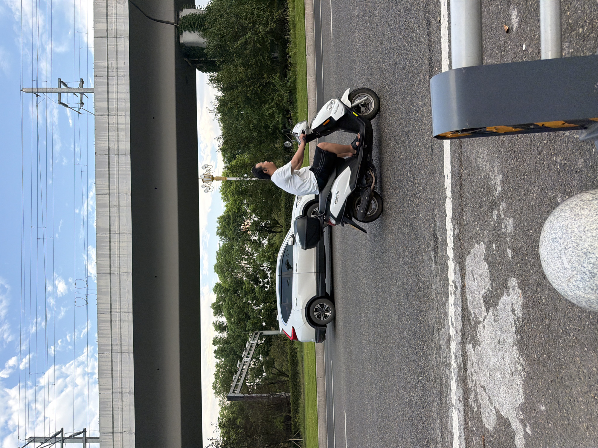 In the city of Jiangjin, China, a man is riding a white scooter on an asphalt road. He is wearing a white t-shirt and black shorts. The scooter has a black storage box on the back. A white SUV is driving in the lane to the left of the scooter. In the background, there are trees and a large concrete overpass with overhead wires. The sky is blue with some white clouds. The image is taken from a slightly low angle, with part of a gray metal guardrail and a spherical concrete object in the foreground.