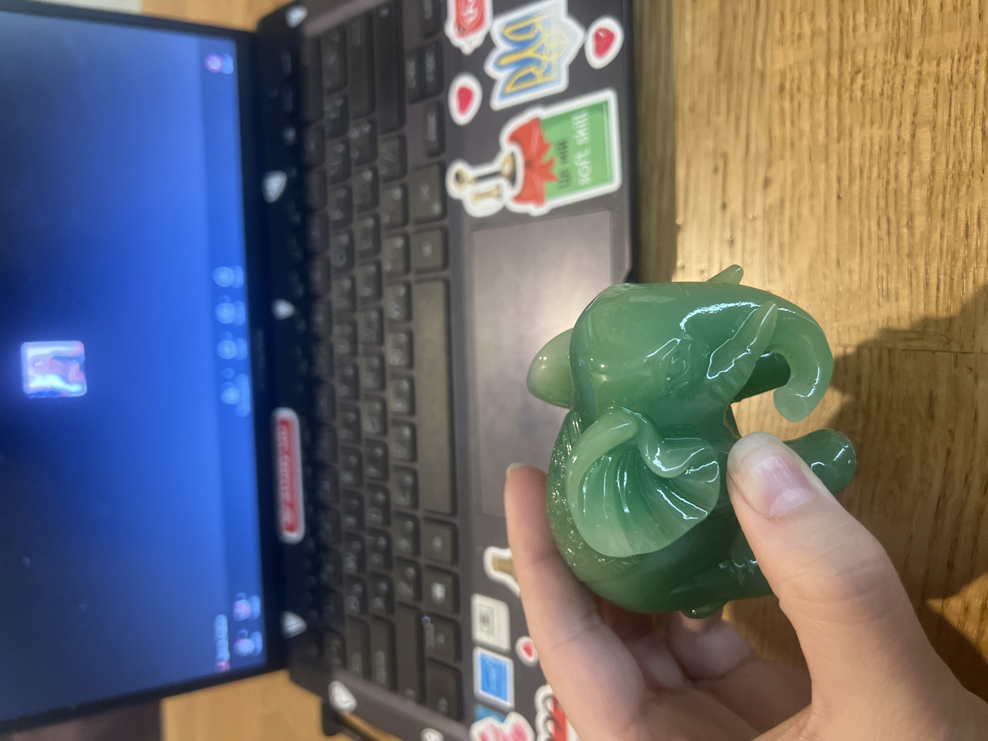 This media file shows a close-up shot of a person's hand holding a small, translucent green jadeite figurine of a dragon. The figurine has detailed wings and a playful expression. The background reveals the keyboard and screen of an open laptop, suggesting a workspace. The laptop is adorned with several stickers, including a Ukrainian trident (Tryzub) and a sticker with text that appears to read "Like You, soft skill". The scene is set on a wooden surface, likely a desk, illuminated by artificial light. The overall mood is casual and domestic. The presence of the Ukrainian trident sticker strongly suggests the location is Kyiv, Ukraine. The time of day and weather are not discernible from the image.