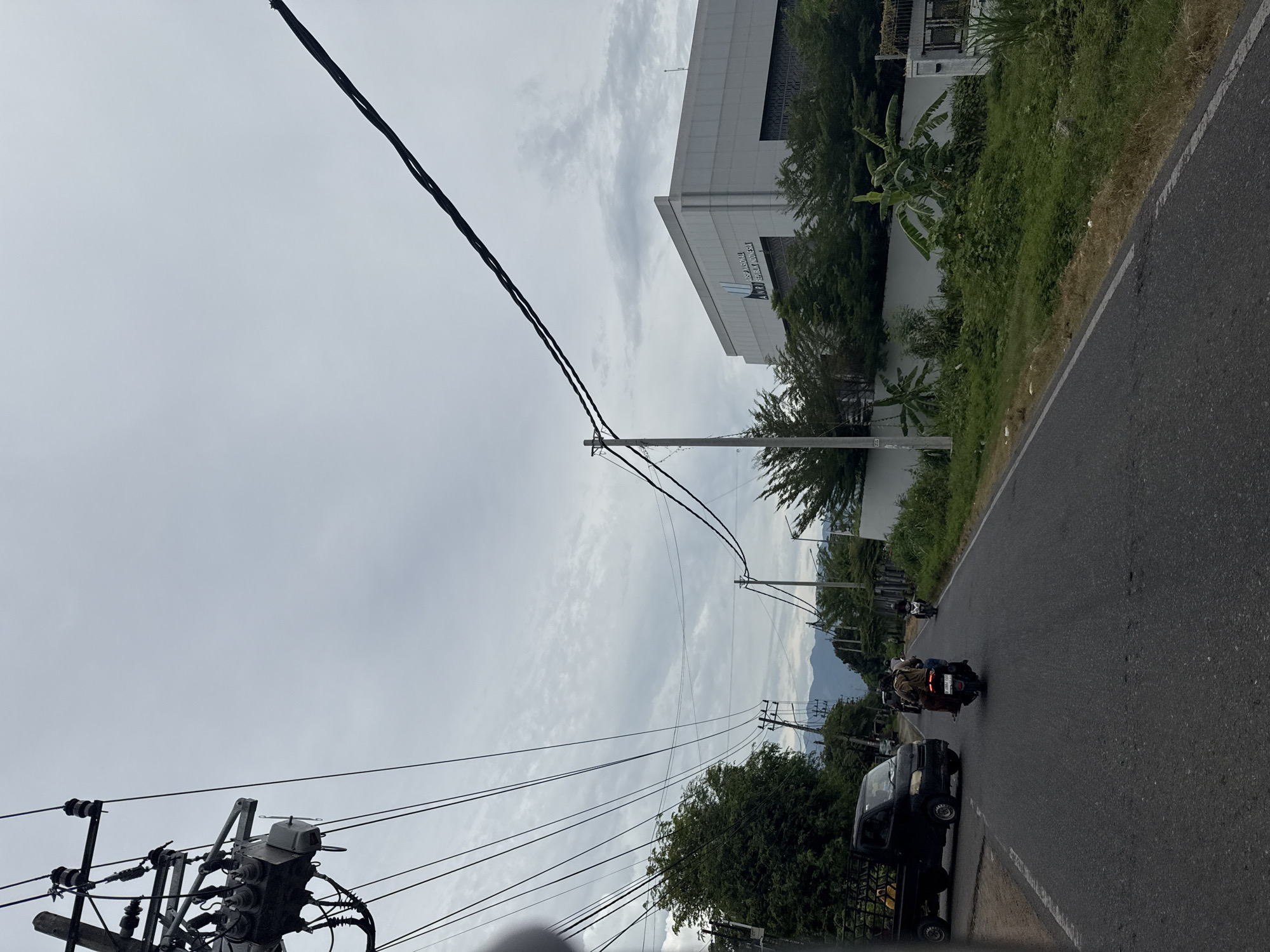 The image depicts a street scene in Manyanglamgarot, Indonesia, under a cloudy sky. A dark-colored pickup truck is parked on the left side of the road. Two individuals are riding a motorcycle in the middle of the road, heading away from the viewer. Further down the road, another motorcycle is visible. On the right side of the road, a modern, light-colored building with the text "ARSIP NASIONAL REPUBLIK INDONESIA" is prominent. Lush green vegetation, including trees and banana plants, surrounds the building and lines the roadside. Electrical poles and numerous wires are visible throughout the scene, indicating an urban or semi-urban environment. The road is paved and appears to be a typical street with white lane markings. The overall impression is a somewhat overcast day in an Indonesian town or city.