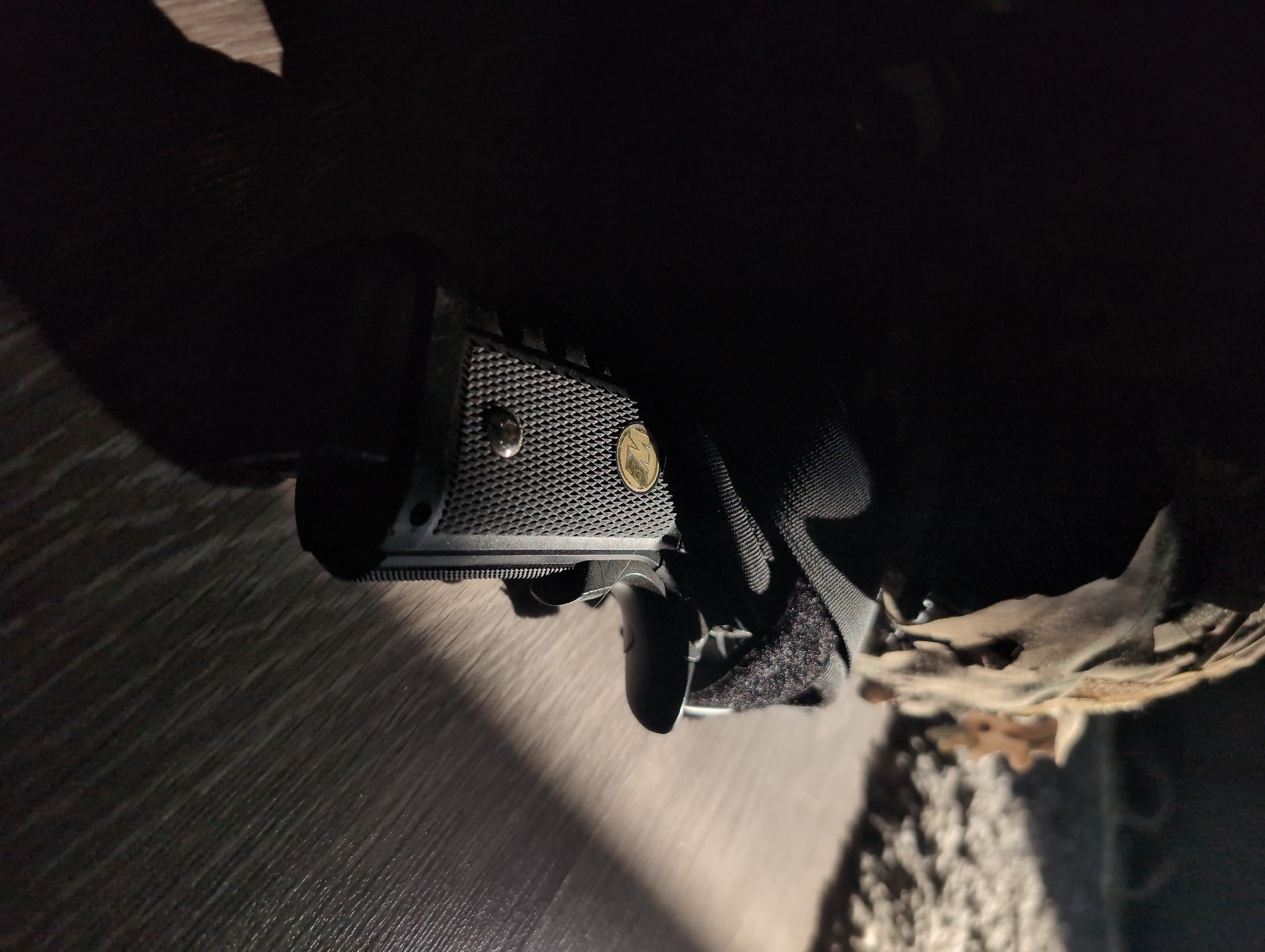 A handgun, secured in a holster, is resting on a wood-patterned surface. The textured grip of the handgun is visible, with a circular emblem embedded in the material. The holster is made of a dark material with a visible Velcro closure. Light shines on the surface next to the gun and holster. The setting is dimly lit, obscuring details of the environment. There is no evidence of the location being Izhevsk, Russia.