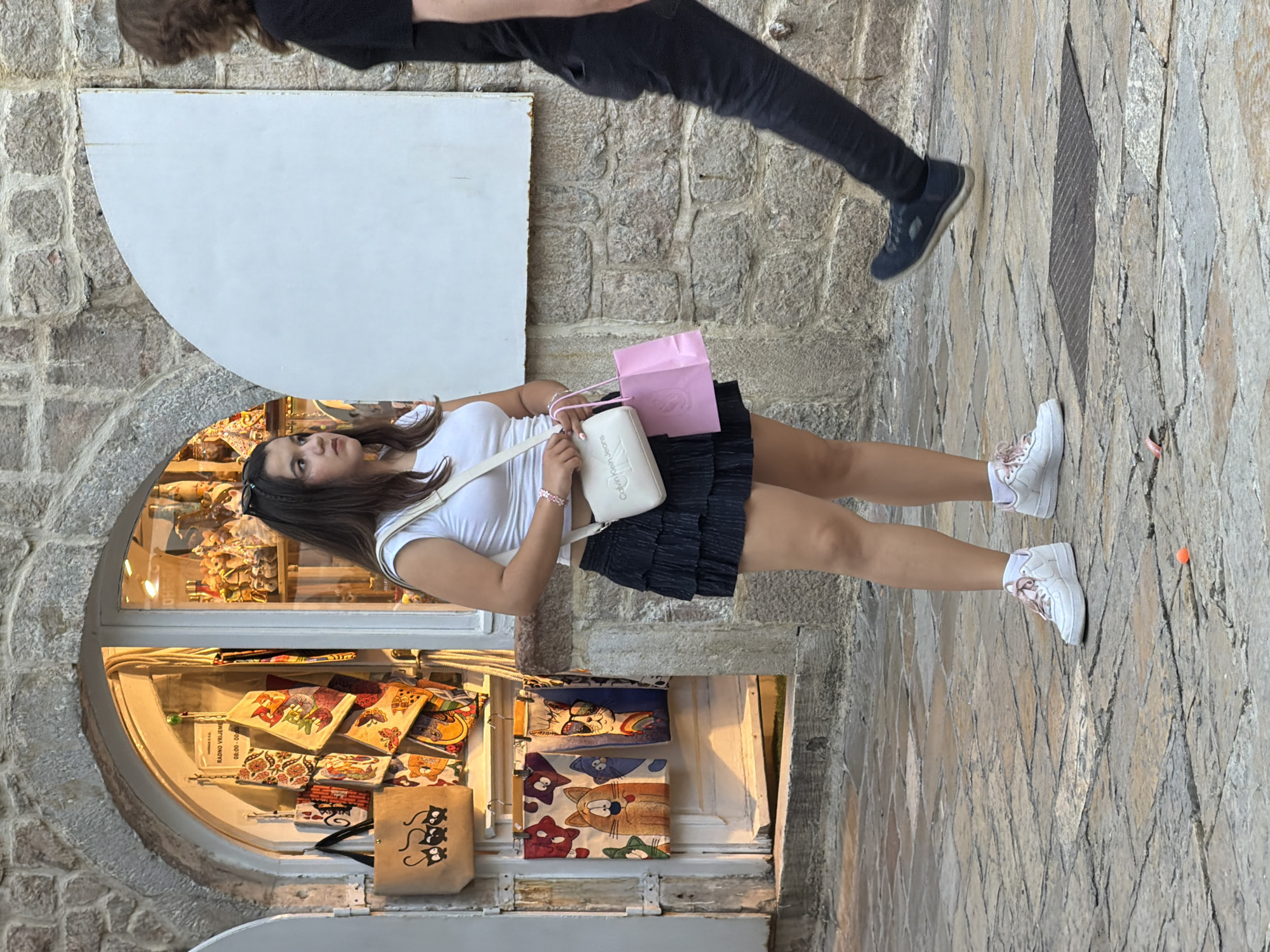 The image depicts a street scene in Budva, Montenegro. A young woman stands facing right, wearing a white crop top, a dark ruffled skirt, and white sneakers with pink laces. She carries a white crossbody bag and a pink shopping bag. Behind her, a shop window displays various souvenirs, including colorful textiles with cat motifs and other items. To the right of the frame, a person wearing a black t-shirt and dark jeans walks past, their legs and lower body visible. The ground is paved with stone. The architecture suggests an older urban environment.