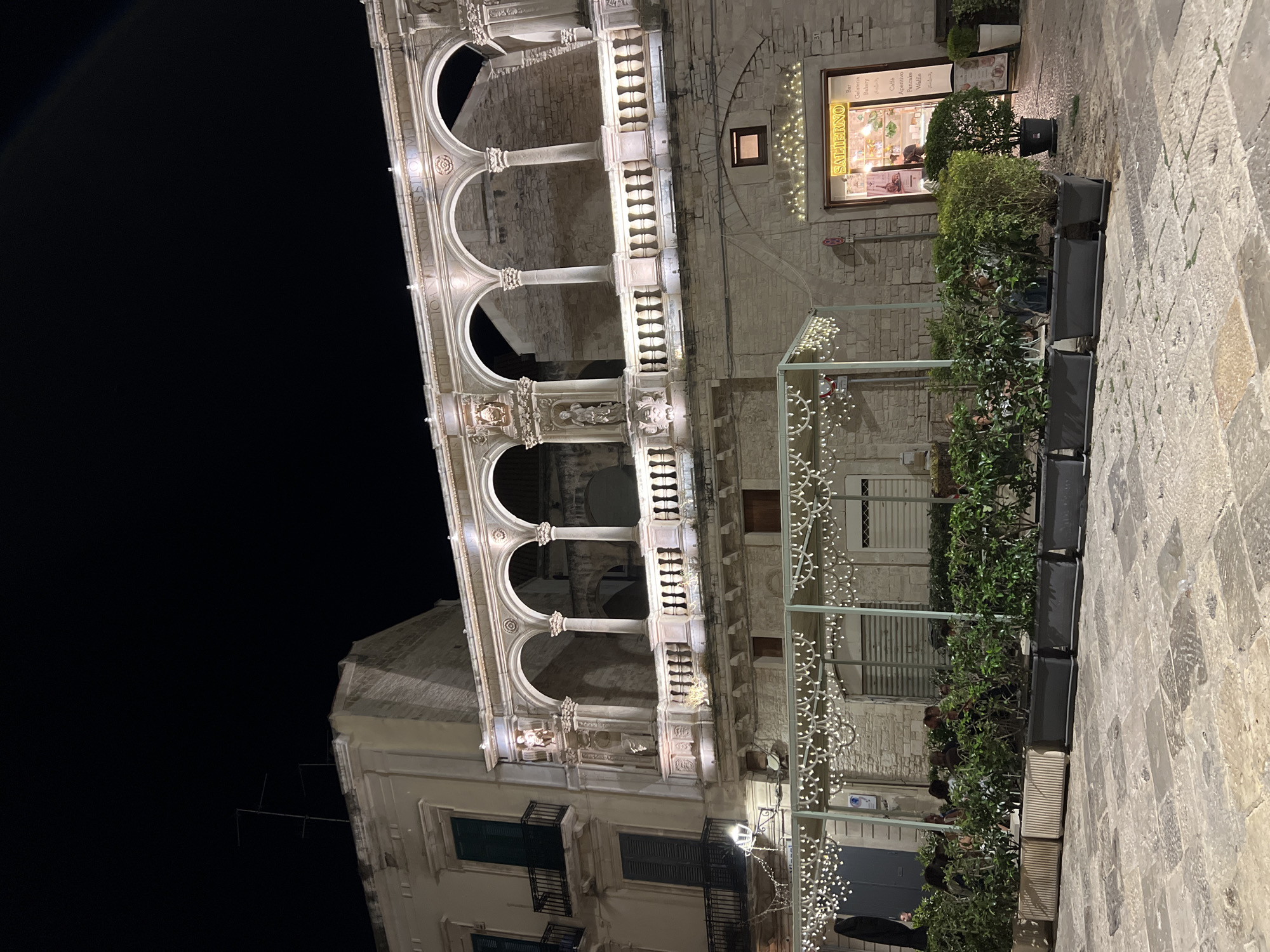 The image shows a nighttime view of a historic building in Bitonto, Italy, featuring a portico with arches and decorative elements. The building is illuminated, highlighting its architectural details. In the foreground, there is an outdoor seating area with tables and chairs, partially obscured by planters filled with greenery. String lights adorn the outdoor seating structure. A sign for "SALIERNO" is visible above a shop entrance, indicating it is a Bar, Gelateria, and Bakery. The paving stones in the square are visible. The sky is dark.