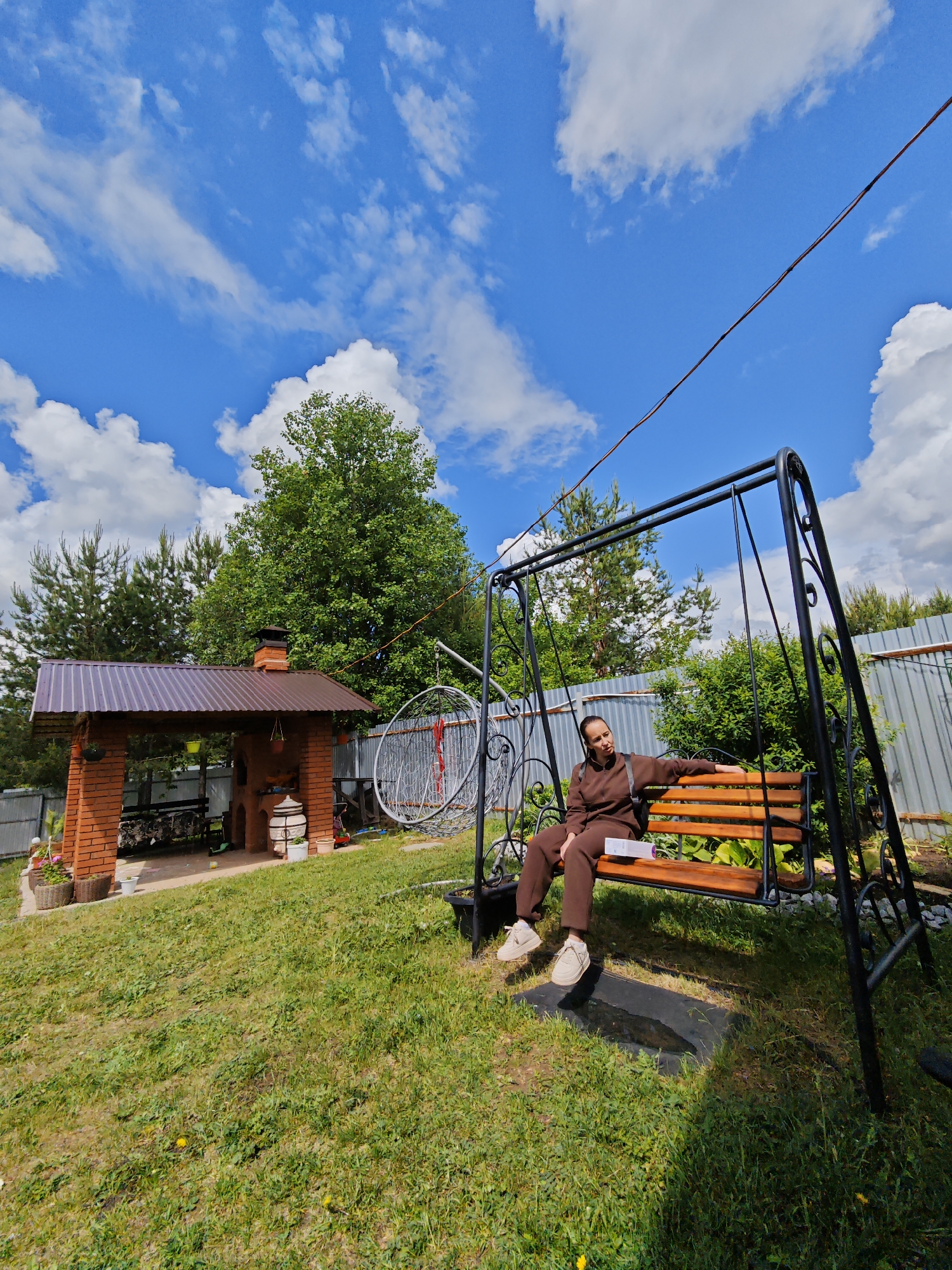 The image presents an outdoor scene, presumably in a backyard or garden. Dominating the foreground is a metal swing set with a wooden bench, upon which a person is seated. This individual wears a brown tracksuit and beige sneakers, and appears to be holding a white rectangular object, possibly a book or a package.
In the background, there's a brick structure that resembles an outdoor cooking area or a small pavilion. It has a brown roof, a brick chimney, and seems to house firewood and other items. To the right of this structure, a decorative silver-colored hoop or frame can be seen.
The overall environment is lush with greenery, including trees and a well-maintained lawn. A fence runs along the edge of the property. The sky is a vibrant blue dotted with fluffy white clouds. Given the context, this scene may be located in a residential area. There is no other information to confirm the location of Markovo, Russia.