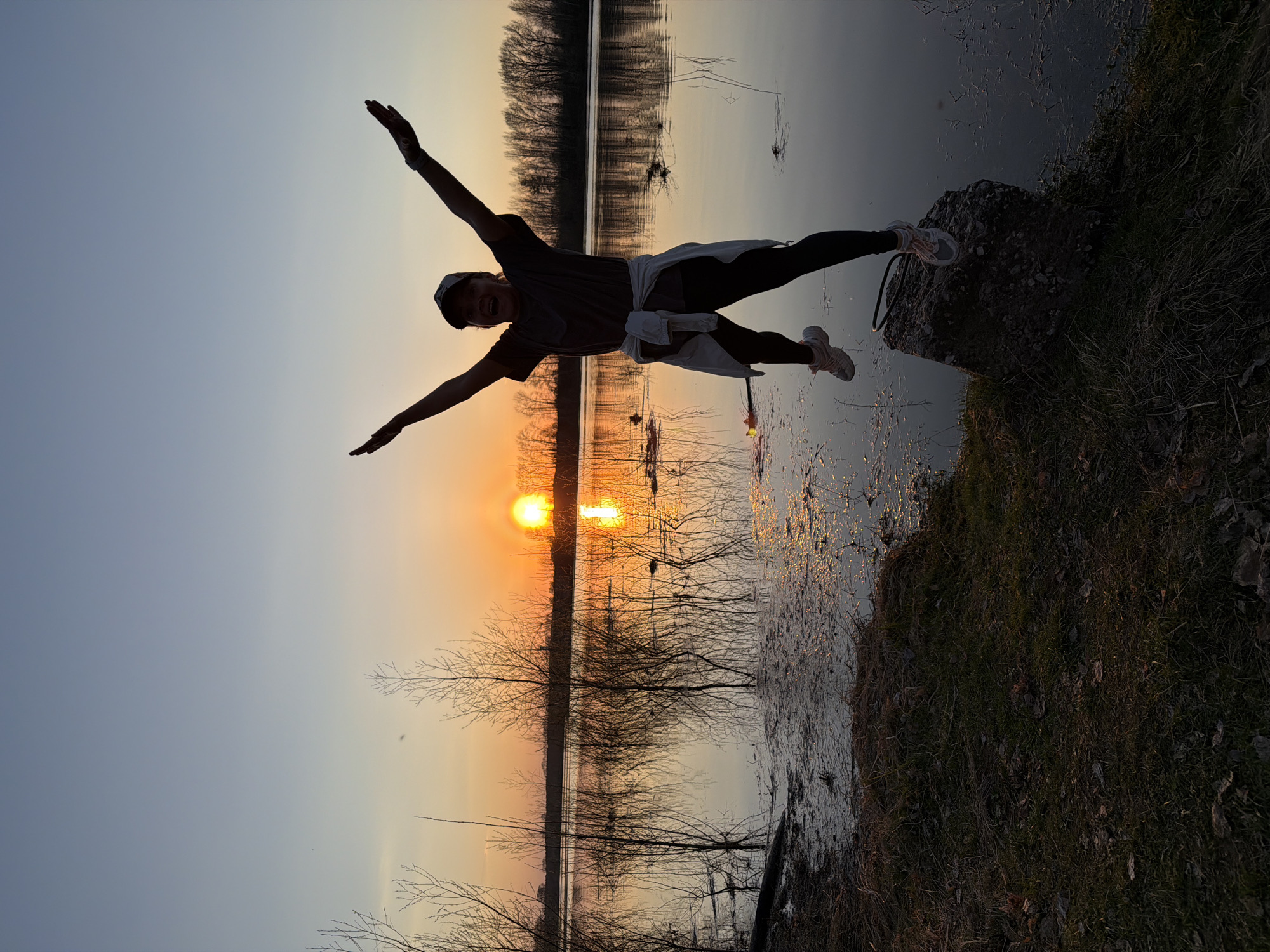 A person is positioned horizontally in the air, with their arms outstretched and legs extended, appearing to be in mid-jump or levitation. They are wearing dark pants, a t-shirt, a white jacket tied around their waist, a baseball cap, and sneakers. The person appears to be over the Kama River, or near the shore in the city of Chaikovsky, Russia. The sun is setting, visible as a bright orb just above a row of bare trees, creating a silhouette effect against the sky. The trees are reflected in the water, and there is vegetation on the right side of the image, next to a body of water.