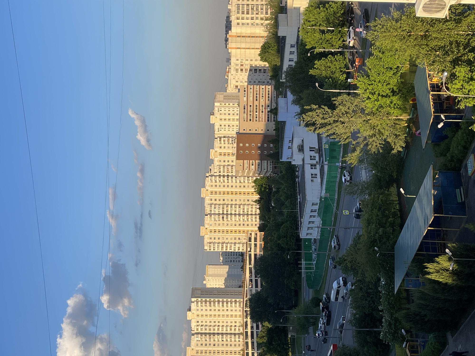 This elevated view captures a residential area in Moscow, Russia, during what appears to be the late afternoon or early evening of a sunny day. The scene is dominated by numerous multi-story apartment buildings, mostly featuring light-colored facades and repetitive window patterns. Many of these buildings are illuminated by warm, golden light, casting long shadows.
A wide street, flanked by lush, mature green trees, runs through the middle ground, suggesting a spring or summer season. Numerous cars are visible on the road, some parked and others in motion. Faint pedestrian crossings can be seen on the asphalt. To the right of the street, a green space or park features additional trees and a children's playground with a distinctive blue-green canopy roof. A large white air conditioning unit is visible on a building rooftop in the upper right.
The sky above is a clear, bright blue with scattered white and grey cumulus clouds, crisscrossed by several thin power lines. A small, indistinct yellow square with an arrow, likely a traffic sign, is visible on a green structure near the street. The overall image depicts a typical, verdant urban environment with ongoing daily activities.