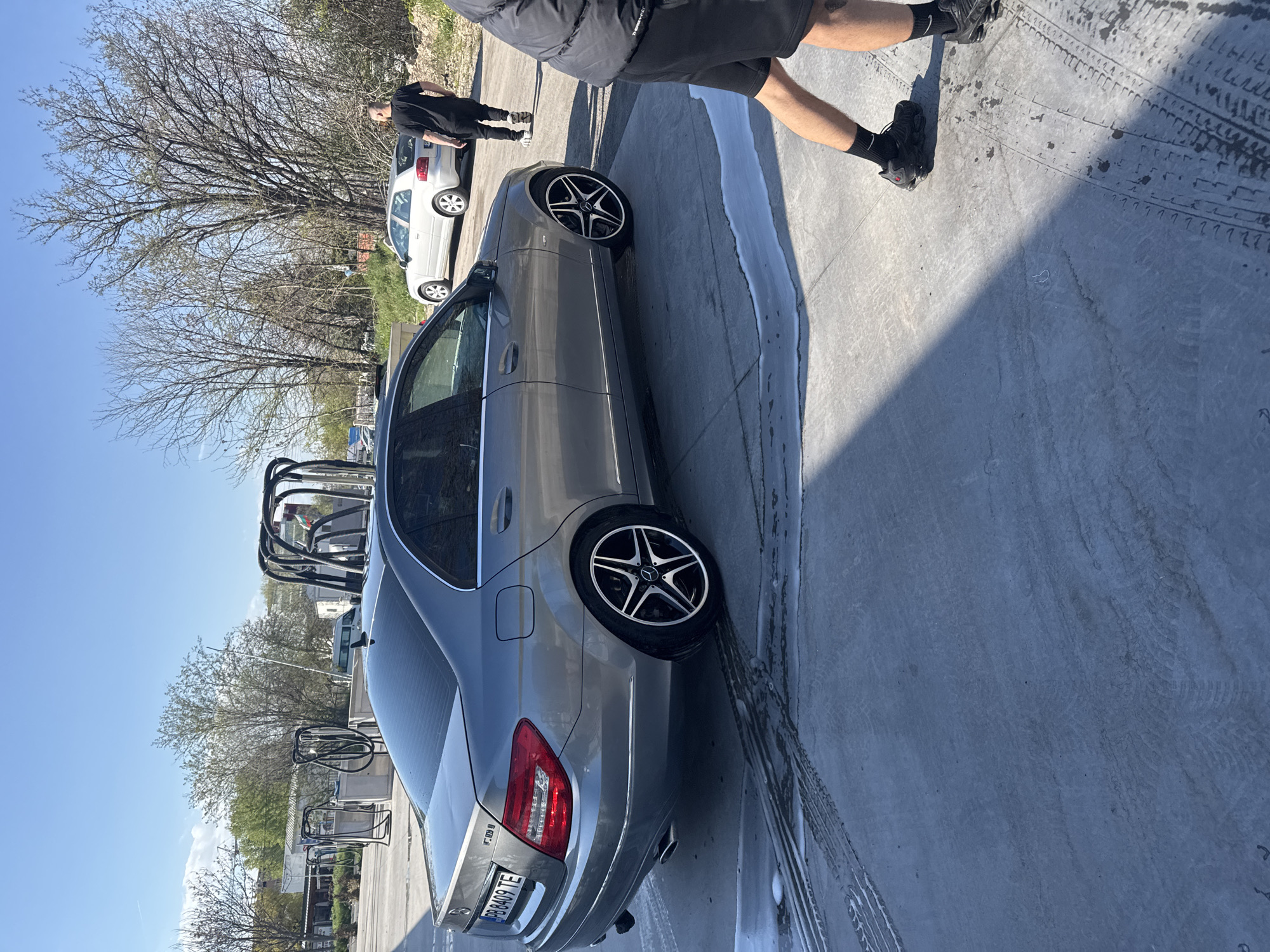The image shows a gray Mercedes-Benz sedan being washed at a self-service car wash. A person wearing a black jacket and shorts is standing near the car. Another individual wearing a black shirt and pants is visible in the background near a white car. The car wash is located outdoors with visible car wash equipment and trees in the background under a blue sky. The license plate of the Mercedes is visible and reads "PB 8409 TE". Given the car plate, the location is likely in Plovdiv, Bulgaria. The concrete surface surrounding the car has tire marks and soap residue, indicating recent washing.