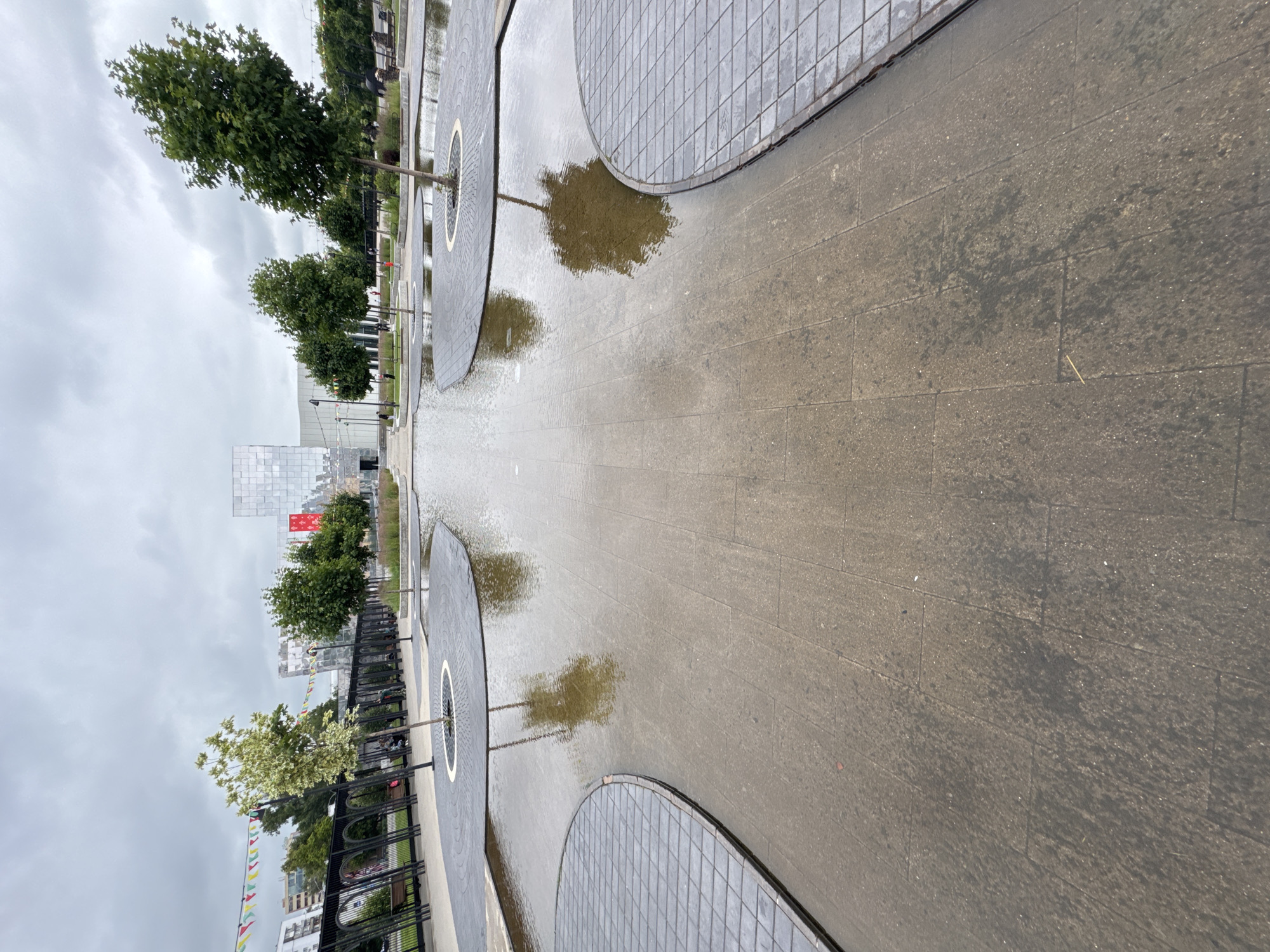 The image depicts a public park area in Nizhny Novgorod, Russia, featuring a large, shallow water feature with reflections of trees. The ground surrounding the water is paved with square tiles, and several circular raised platforms with trees are situated within the water. In the background, there is a modern, glass-covered building with a red banner, flanked by trees and a black metal archway structure. The sky is overcast and cloudy. A few people are visible in the distance, walking or sitting on benches. Decorative flags are strung across the area.