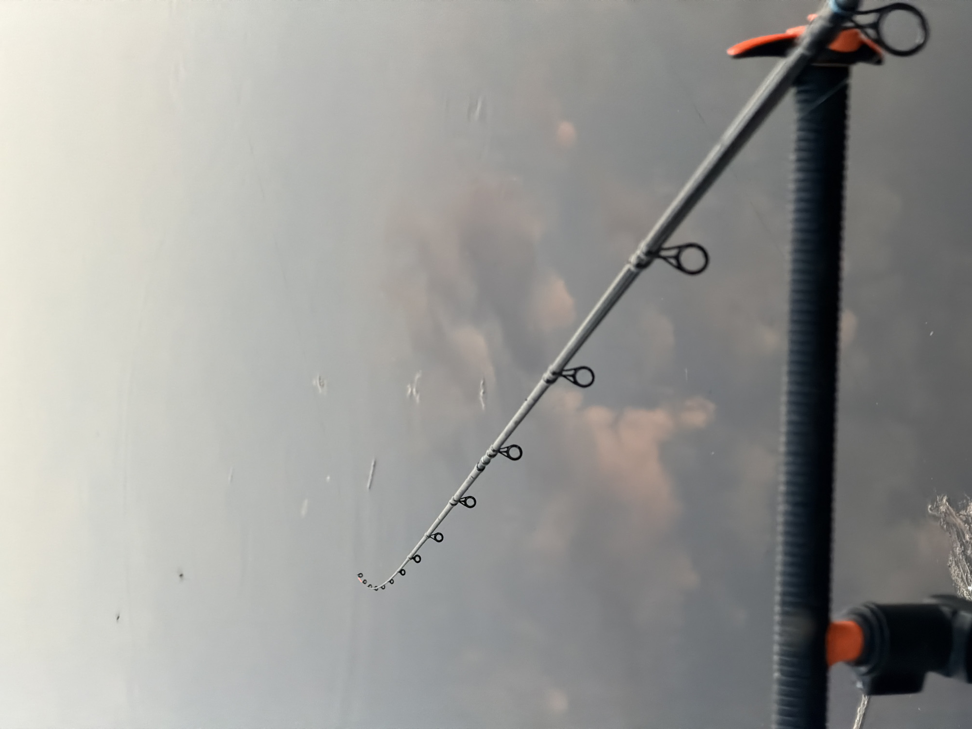 The image shows a fishing rod bent downwards, with a cloudy sky visible in the background. The rod is grey with black guides and an orange accent near the reel seat, which is mounted on a black, textured vertical support. The rod tip is heavily bent, indicating tension on the line. The background sky appears overcast and possibly reflecting off a surface, blurring any specific details. The location is not discernible from the image, and there is no information about the city of Chaykovsky, Russia.