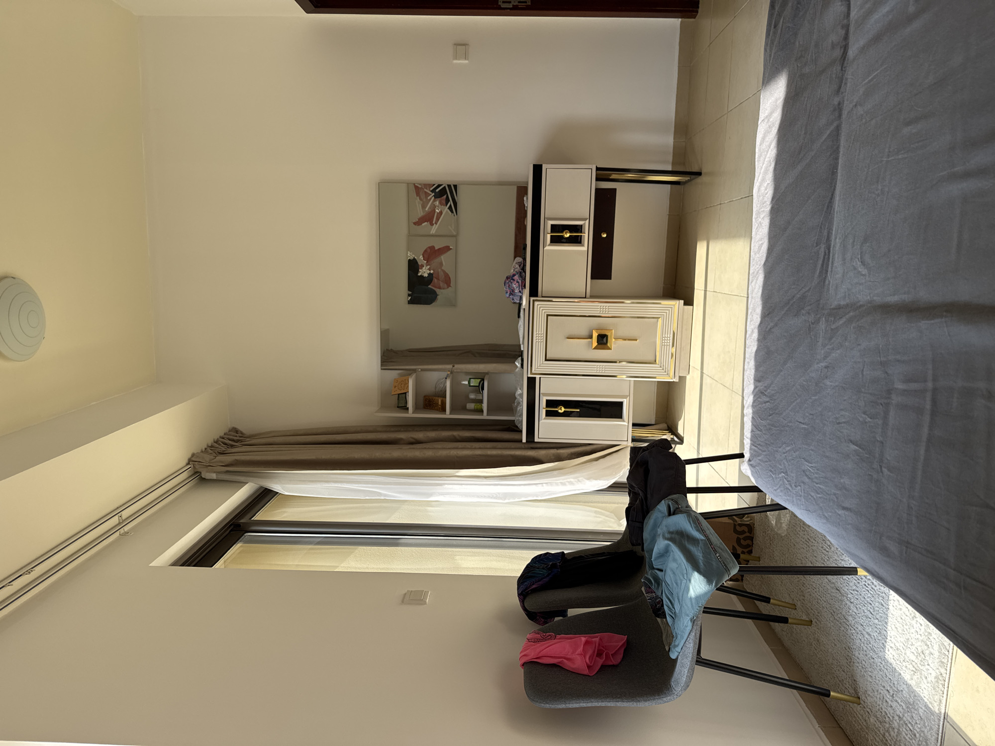 The image depicts a bedroom scene, likely within a modern apartment in Dubai, United Arab Emirates. A large window with sheer and opaque curtains lets in natural light, casting shadows across the tiled floor and a textured rug. Two grey chairs with black legs tipped with gold are positioned near the window. These chairs are draped with clothing, including a pink top and blue jeans.
In the background, a vanity table with a large mirror stands against the wall. The vanity has a white and gold ornate design, featuring drawers and a central cabinet with a prominent gold lock-like embellishment. The mirror reflects two abstract artworks on the wall. To the left of the vanity, a tall, slim shelving unit holds various items, possibly toiletries or decorative objects. A ceiling light fixture is visible in the upper portion of the image. To the right, a dark wooden door frame is partially visible, suggesting another room or the entrance to the bedroom. The overall impression is of a tidy but lived-in space, with personal items casually placed.