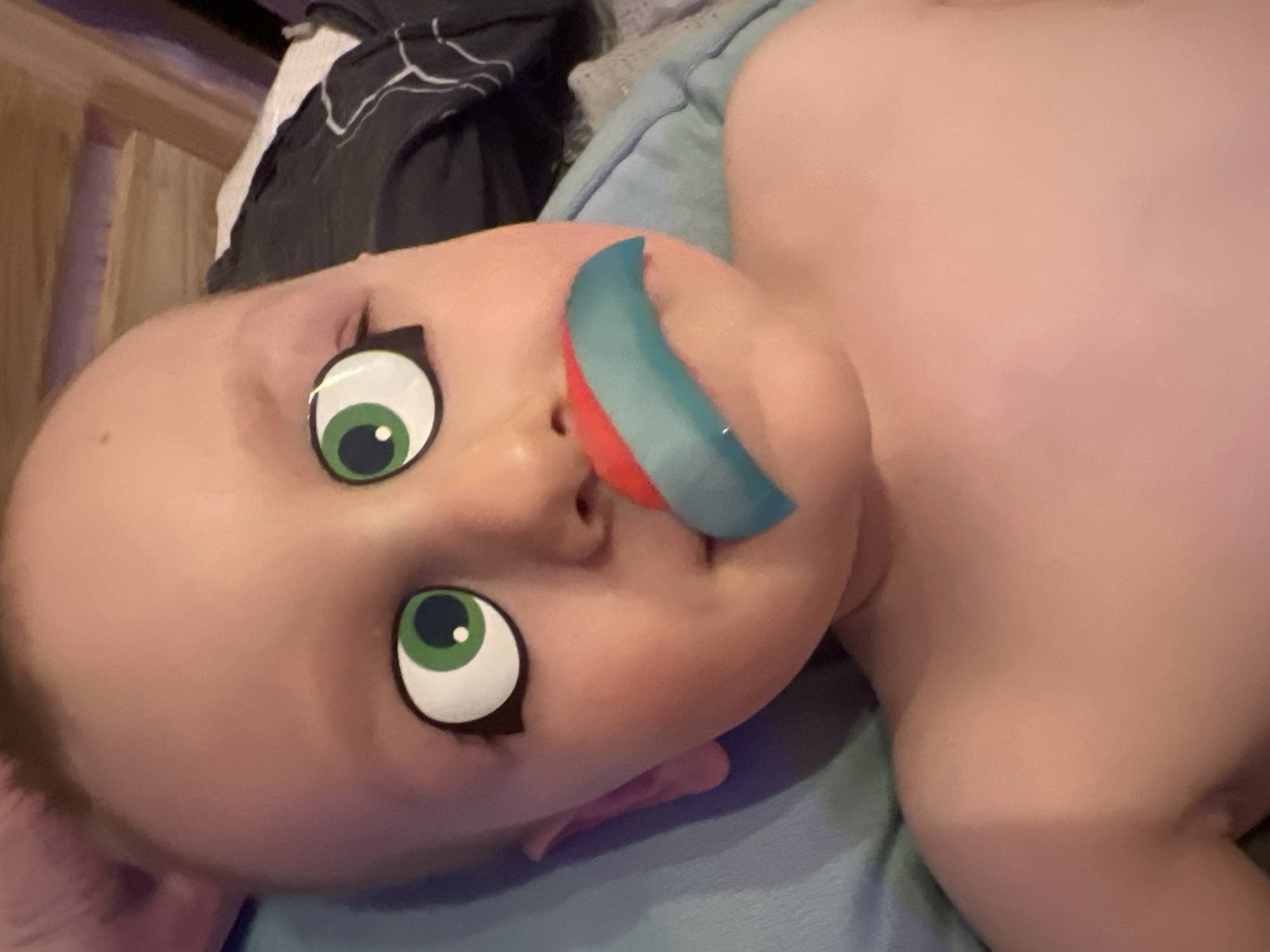 A baby is lying on a light blue blanket. The baby has digitally-imposed cartoon eyes and a blue and red object is digitally placed over their mouth. Bedding and a wooden headboard are visible in the background. No location is discernible in this image.