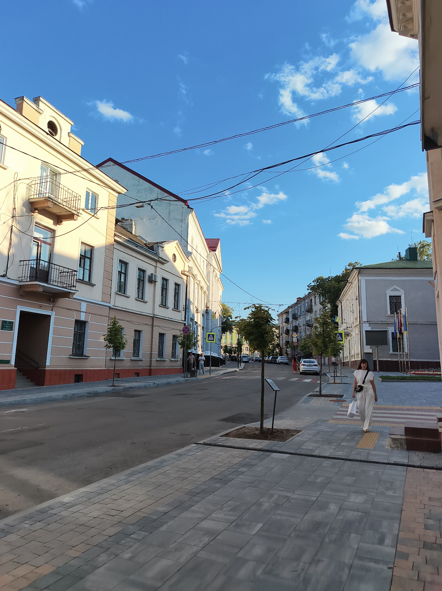 The image depicts a street scene in Ternopil, Ukraine, likely during daylight hours given the bright sky and shadows. The perspective is from a sidewalk looking down a street lined with buildings and trees.
On the left, a pale yellow building with red trim and balconies dominates the foreground. Adjacent to it, a lighter colored building with a red roof extends further down the street. Several windows, some with air conditioning units, are visible on the building facades. Decorative architectural elements, such as cornices, are present.
The street itself is paved with asphalt and appears to be a two-way road with parked cars visible in the distance. On the right side of the street, a woman in a white outfit and a crossbody bag is walking away from the camera, carrying a white bag. She is on a modern pedestrian walkway paved with grey and beige square tiles, bordered by a strip of yellow tactile paving. Further down the street, other pedestrians, traffic signs, and flags are visible.
The sky is a vibrant blue with scattered white clouds. The overall impression is of a well-maintained urban environment.
