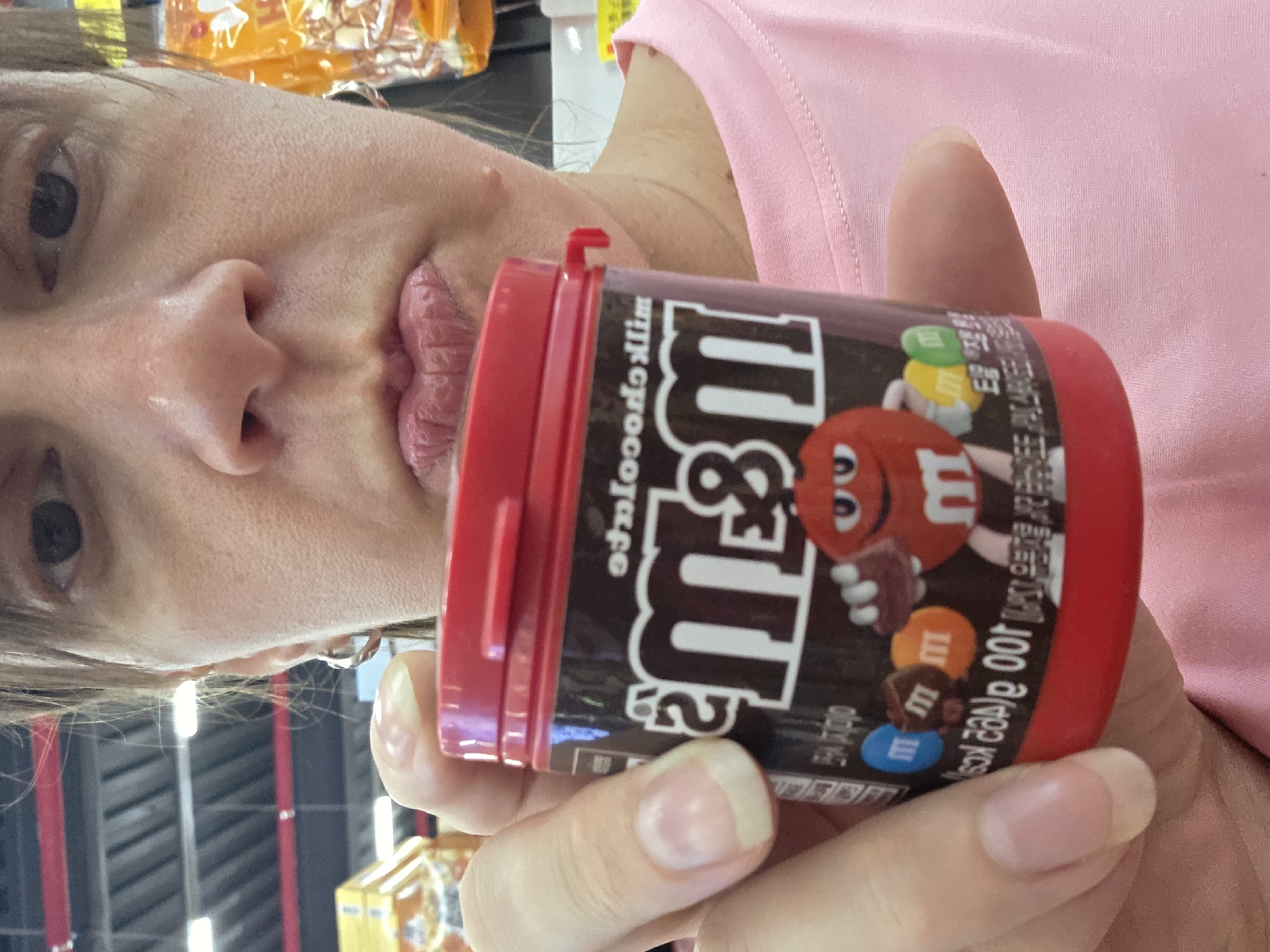 A woman's face is framed by the top of the image, with the lower portion of her face and a container of M&Ms in the foreground. The woman has brown hair and is wearing a pink shirt. She is holding the red-lidded container of M&Ms in her right hand, bringing it closer to the camera. The M&Ms container is dark brown with the M&M logo prominently displayed. The background is blurred, showing what appears to be a grocery store or retail environment, with shelves and lighting visible. The scene is likely in Dunnae-myeon, South Korea, given the context.