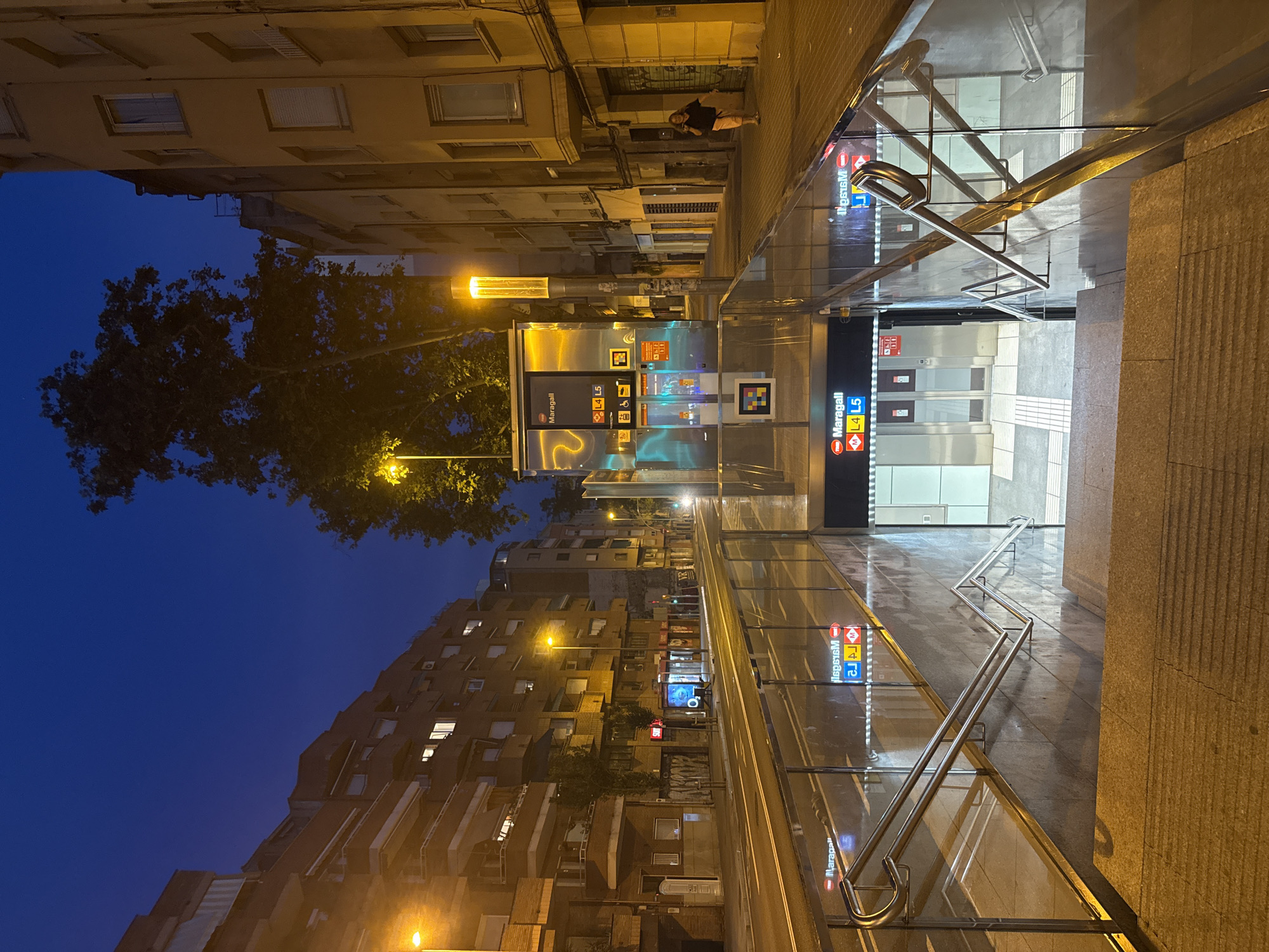 This is a nighttime, low-angle shot of the entrance to the Maragall metro station in Barcelona, Spain. The scene is illuminated by streetlights and the lights from the station itself. In the foreground, a set of concrete stairs descends into the metro station, bordered by glass and metal railings. The station entrance is visible below, with signs indicating "Maragall" and "L4 L5". Across the street, buildings line the road, with some windows lit. A large tree stands to the left of the metro entrance, its branches silhouetted against the dark blue night sky. A single woman is visible on the sidewalk to the right, walking away from the camera. The street appears to be a tram line, with tracks running down the center.