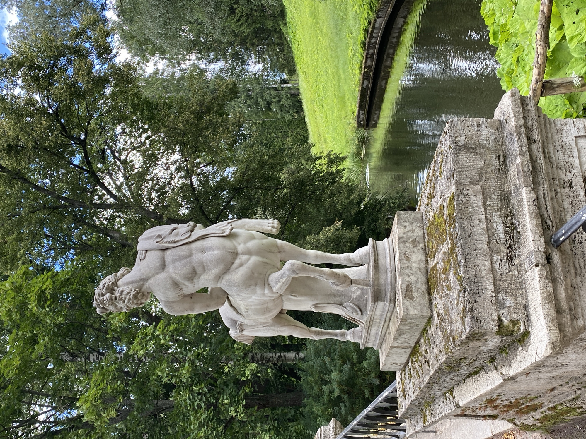 The image depicts a statue of a centaur in the city of Pavlovsk, Russia, situated in a park that appears to be part of the Pavlovsk Palace complex. The statue, carved from white stone, is positioned on a multi-tiered stone pedestal, which is weathered and partially covered in moss. The centaur statue faces away from the viewer, showcasing its muscular back and equine hindquarters.
The surrounding environment is lush and verdant, characteristic of a park or garden setting. A narrow, calm body of water, possibly a canal or river, flows alongside a grassy incline. The water's surface reflects the trees and sky, creating a sense of depth and tranquility. Dense green foliage, including mature trees with full canopies and smaller bushes, frames the scene. The sky above is partly cloudy, with patches of blue visible through the leaves. A black metal railing is visible on the left side of the image, suggesting it may be part of a bridge or walkway. The overall atmosphere is serene and picturesque, highlighting the artistry of the statue and the natural beauty of its surroundings.