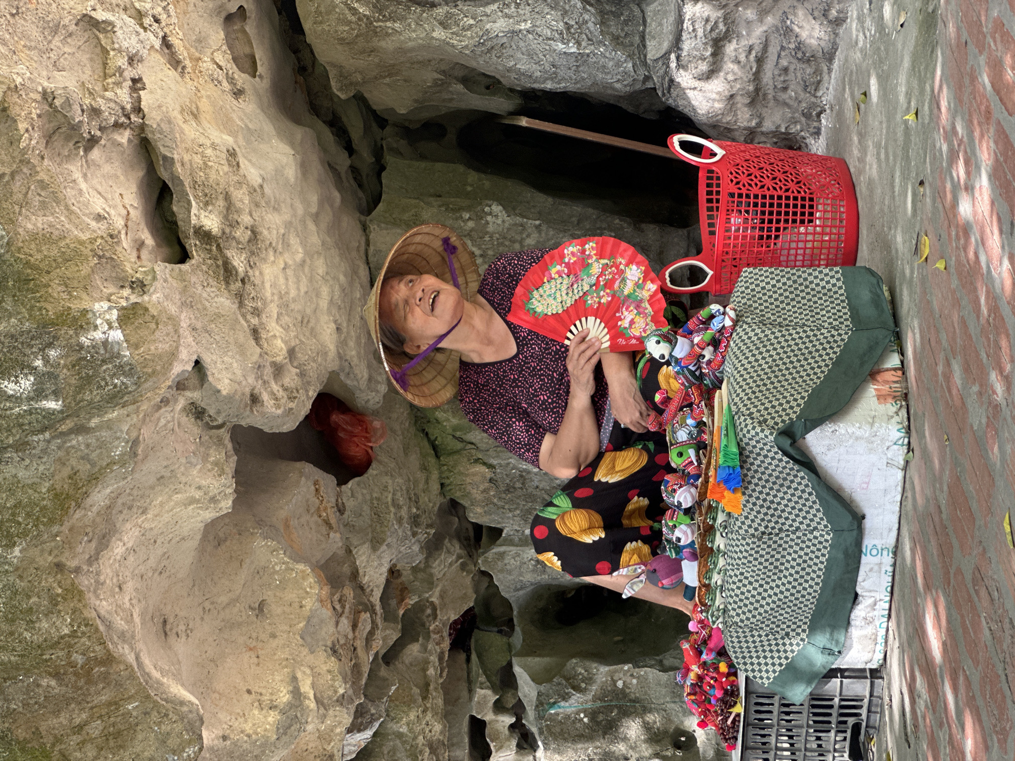 A woman wearing a conical hat and a patterned dress sits in a cave, fanning herself with a red fan. She is surrounded by various small, colorful trinkets and toys displayed on a table covered with a green and white checkered cloth. A red basket with handles is placed to her right. The cave walls, composed of natural rock formations, form the backdrop of the scene, with some greenery visible on the upper surfaces. The ground appears to be made of brick or tile. The image captures a moment of a vendor selling goods in a natural, sheltered environment.