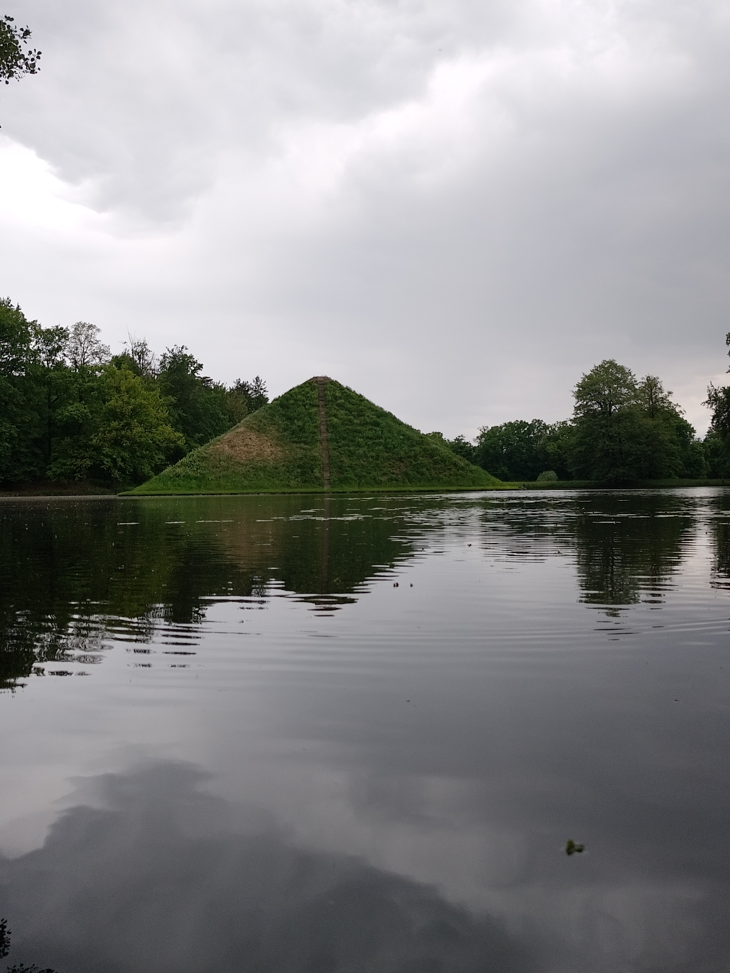 A view of a pyramid-shaped landmass in a body of water. The pyramid is grass-covered with a visible path leading to the top. Trees surround the water body. The sky is overcast. The water's surface shows ripples and reflections of the sky and surrounding elements. The location is likely the Pyramid of Austerlitz in Woudenberg, Netherlands.
