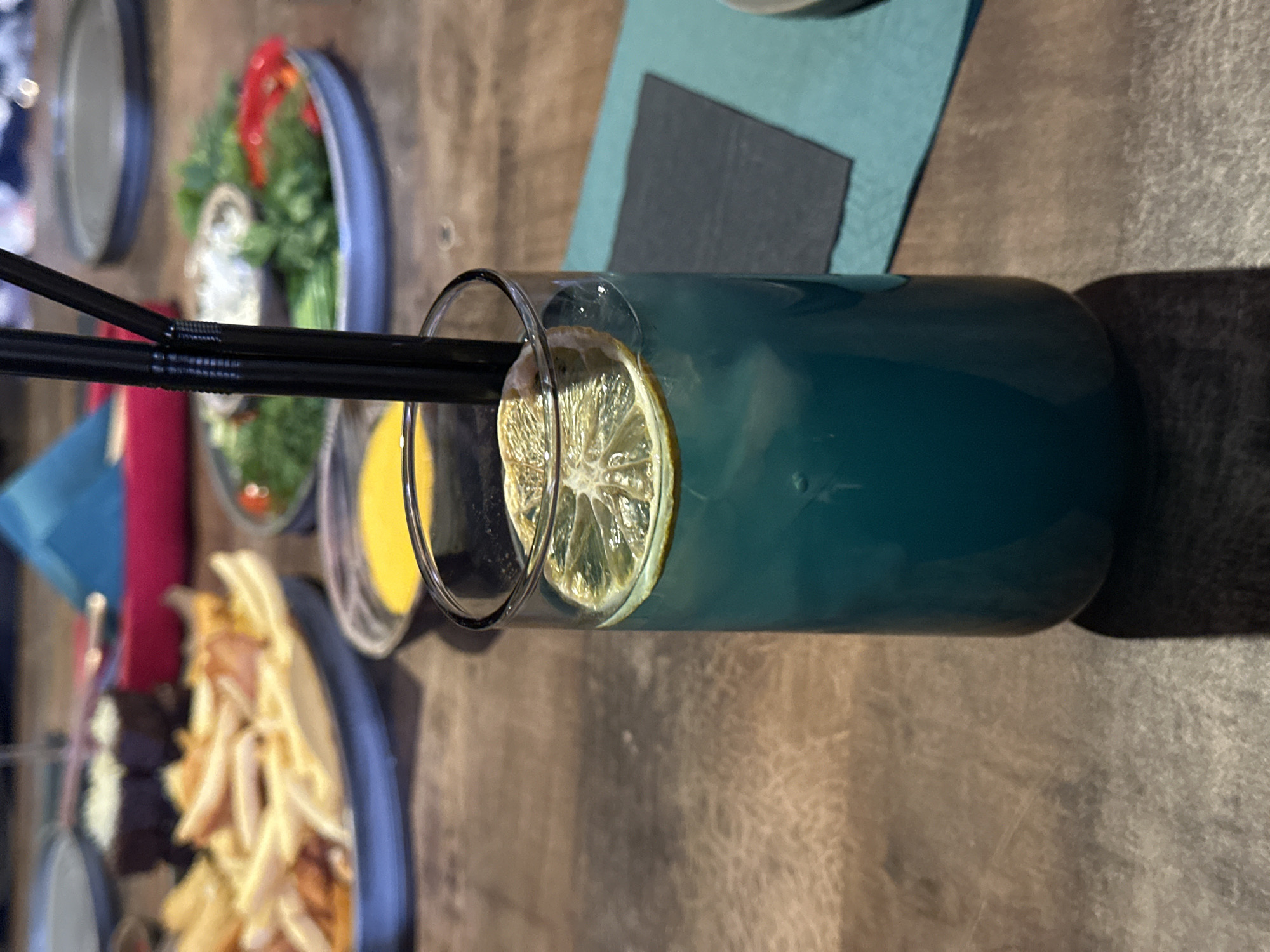 A close-up, slightly rotated, eye-level shot depicts a tall, cylindrical glass containing a blue-green liquid, garnished with a lemon slice and two black straws. The glass rests on a light-brown, textured surface. Adjacent to the glass are several plates holding food items, including what appears to be salad, fries, and small appetizers. The location is Chaikovsky, Russia.