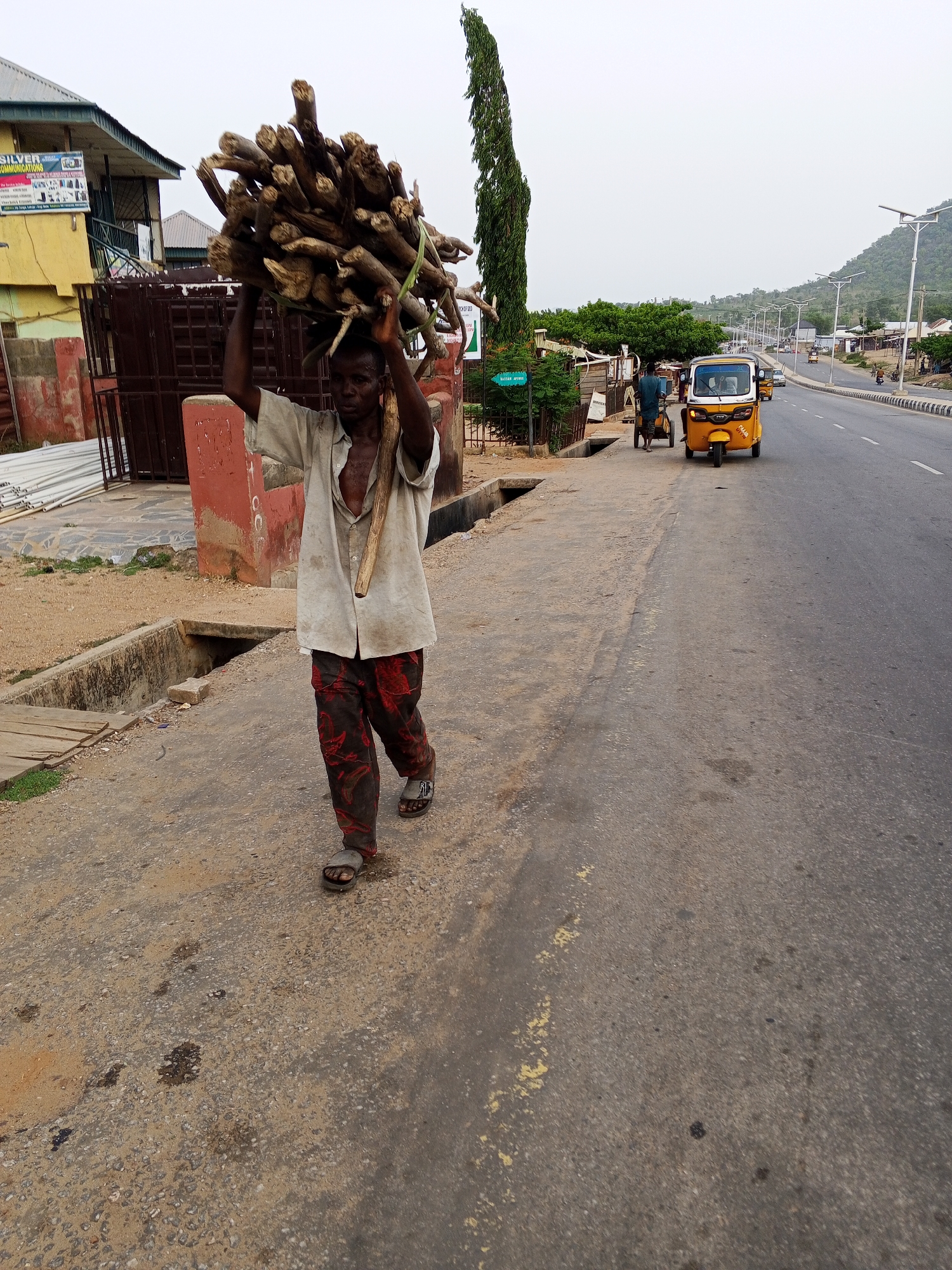 The image captures a street scene in Zango-ndaji, Nigeria, featuring a man carrying a substantial bundle of firewood on his head. The man, who has a dark complexion, walks on a dirt and gravel path adjacent to a paved road. His arms are raised to steady the large, irregularly shaped stack of wood, which is bound together by a visible green cord. He is dressed in a light-colored, unbuttoned collared shirt and dark trousers with a distinct reddish-orange pattern, along with dark sandals.
To the man's left, there is a building with a yellow upper facade and a dark green roof, displaying a sign that reads "SILVER COMMUNICATIONS" at the top, and further down, smaller text includes "SERVICES" and "HAJJ AND UMRAH." Nearby are metal gates and a pile of white pipes, suggesting construction or storage. A concrete drainage channel runs along the path, partially covered by a wooden plank. A small green sign is partially visible, showing "BATAN SPRIN."
To the man's right, a paved two-lane road stretches into the distance, marked by a yellow and black striped curb. A yellow auto-rickshaw (tuk-tuk) is visible on the road further down, and another person is walking near it. The road is lined with modern streetlights. In the background, rolling hills rise under an overcast sky, with scattered buildings and trees dotting the landscape. A tall, slender evergreen tree stands prominently in the mid-ground. Another sign in the background reads "BACK OF 2020."
The scene depicts a common daily activity of transporting goods manually in a semi-urban setting, under natural daylight conditions.