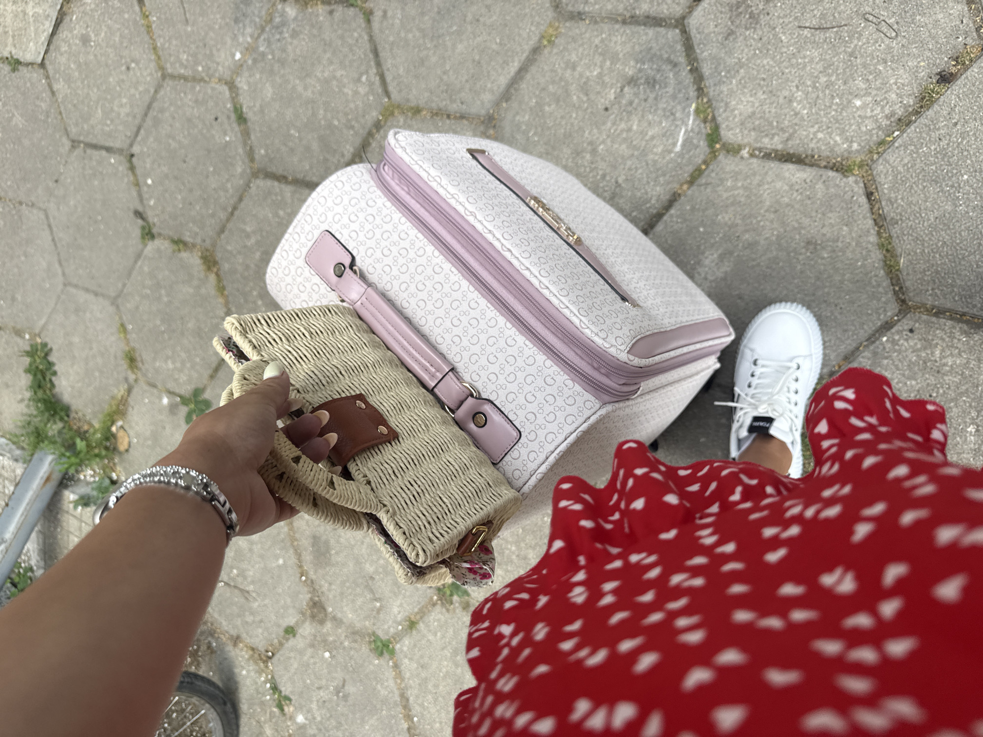 The image shows a woman's perspective looking down at her luggage and attire as she stands on a hexagonal paved pathway, possibly in Plovdiv, Bulgaria. She is holding a small straw purse in her left hand, which is adorned with a silver bracelet. Beside her, on the pathway, are two bags: a larger white and pink patterned travel bag with the "Guess" logo visible, and the straw purse she is holding. She is wearing a vibrant red dress with a white polka dot pattern and white sneakers. The scene suggests she is preparing to travel or has just arrived somewhere. The lighting indicates it is daytime, and the presence of greenery and the outdoor setting suggest fair weather. The visible text "Guess" is printed on the larger bag.