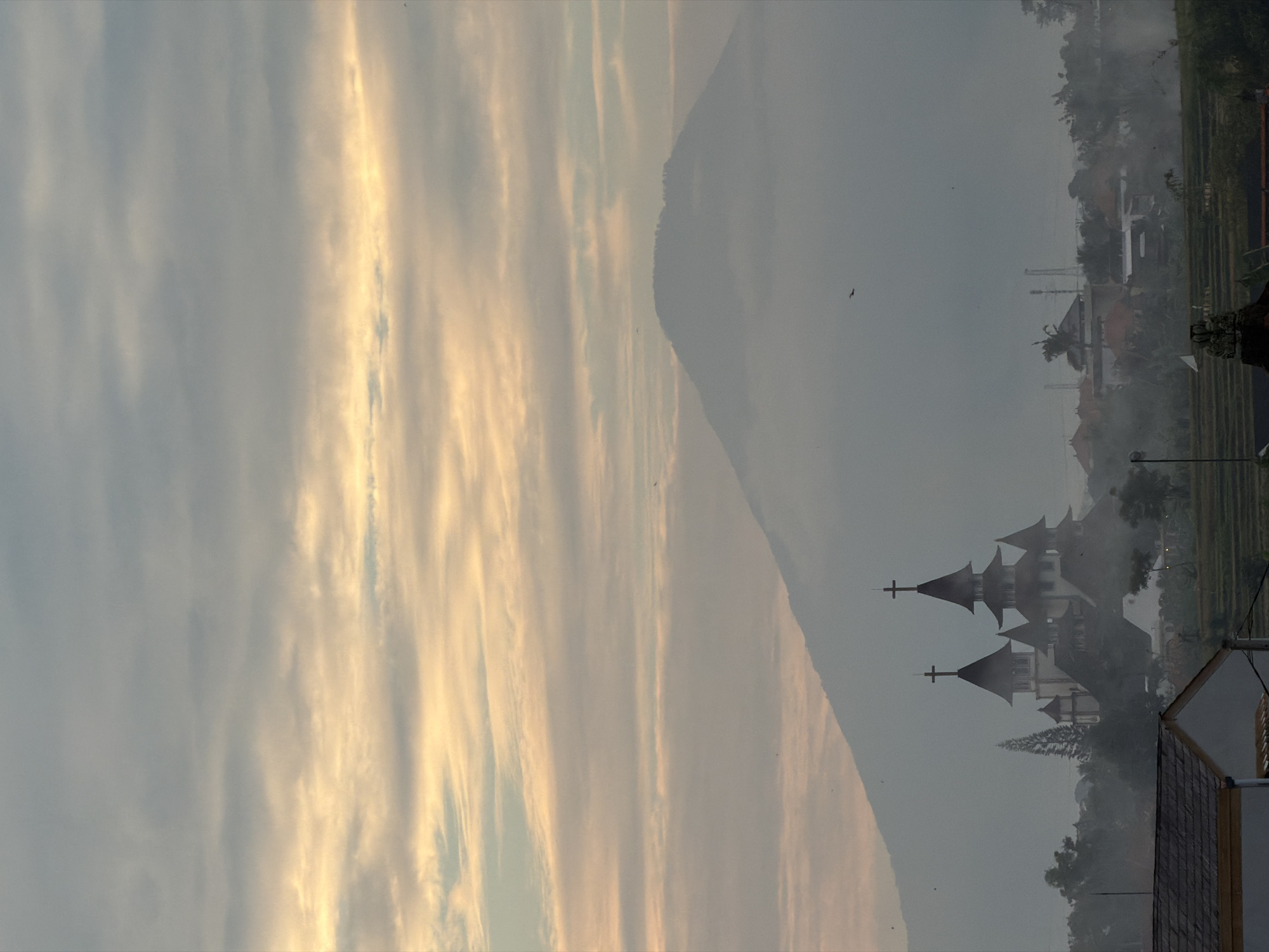 The image depicts a serene landscape at dawn or dusk, with a large, misty mountain dominating the background. In the mid-ground, a prominent church with multiple spires and crosses stands amidst a cluster of buildings and lush greenery. The sky above is a soft gradient of pale blue and warm yellow, striated with thin clouds, suggesting the early or late stages of daylight. The foreground reveals a portion of a building with a tiled roof, and further back, terraced fields suggest a rural or semi-rural setting. The overall atmosphere is tranquil and slightly hazy, with the mist softening the edges of the landscape.