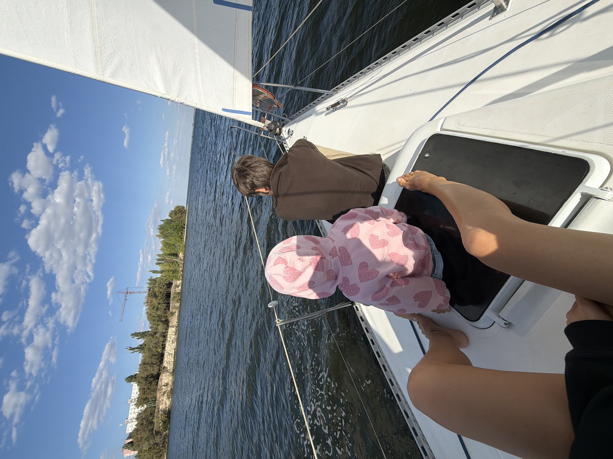 Two people, one an adult male and one a young child wearing a pink hooded sweatshirt with a heart pattern, are seated on the deck of a white sailboat. The adult male is wearing a brown shirt and beige pants, and is facing away from the camera. The child is facing away from the camera and to the left, looking towards the water. A third person, likely an adult, is partially visible in the foreground, with their legs extended and resting on the deck. The sailboat is on a body of water, likely a river or lake. In the background, a shoreline is visible with trees and a building. A construction crane is also visible on the distant shore. The sky is blue with scattered white clouds. The scene appears to be taking place during the daytime, with bright sunlight. The location is Kamyshin, Russia, on the Volga River.