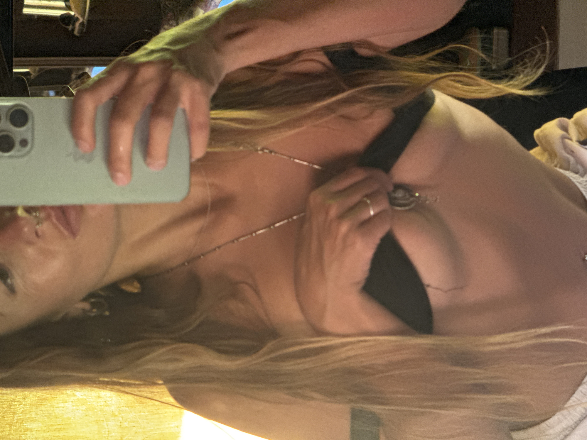 A person is taking a selfie in a room, holding a light blue smartphone in their right hand and using their left hand to adjust a black garment on their chest. The person has long, blonde hair and is wearing a necklace with a pendant. They are also wearing rings on their fingers. The background is dimly lit, with a yellow curtain on the left and what appears to be a mirror or reflective surface on the right, showing some shelves or furniture. The lighting is warm and appears to be coming from the left, casting a glow on the person's skin and hair.