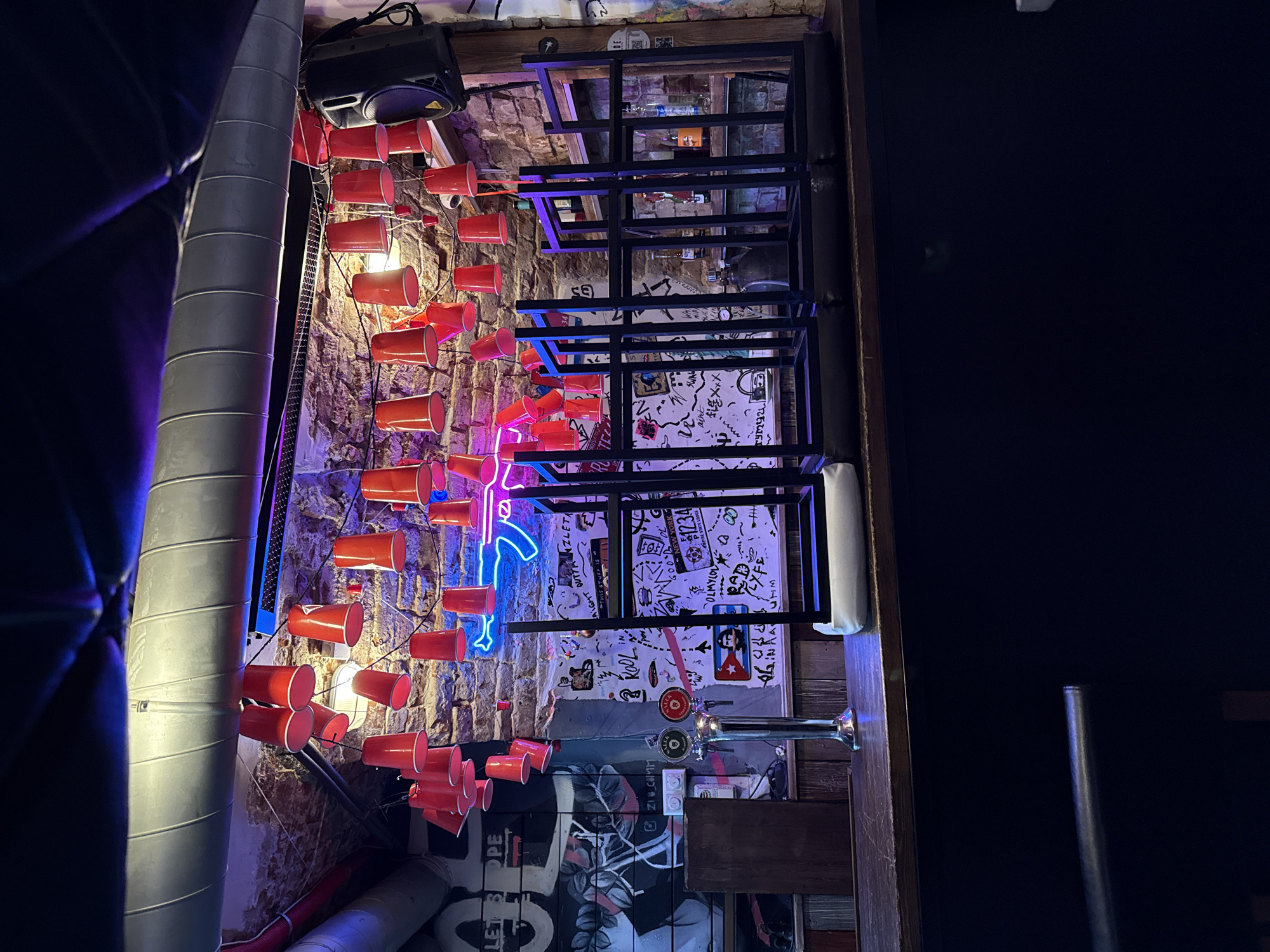 The image depicts an interior space with a brick wall adorned with red plastic cups connected by wires and illuminated by a neon sign shaped like a figure running or jumping. A black metal shelving unit stands against the wall, containing various objects and displaying wall art or posters. A beer tap system is affixed to the wall below the shelving unit, along with graffiti and stickers. To the left, there's a silver ventilation pipe. The scene has a dark, bar-like atmosphere.
Based on the available information, it is impossible to determine if this image is from Krasnoyarsk, Russia.