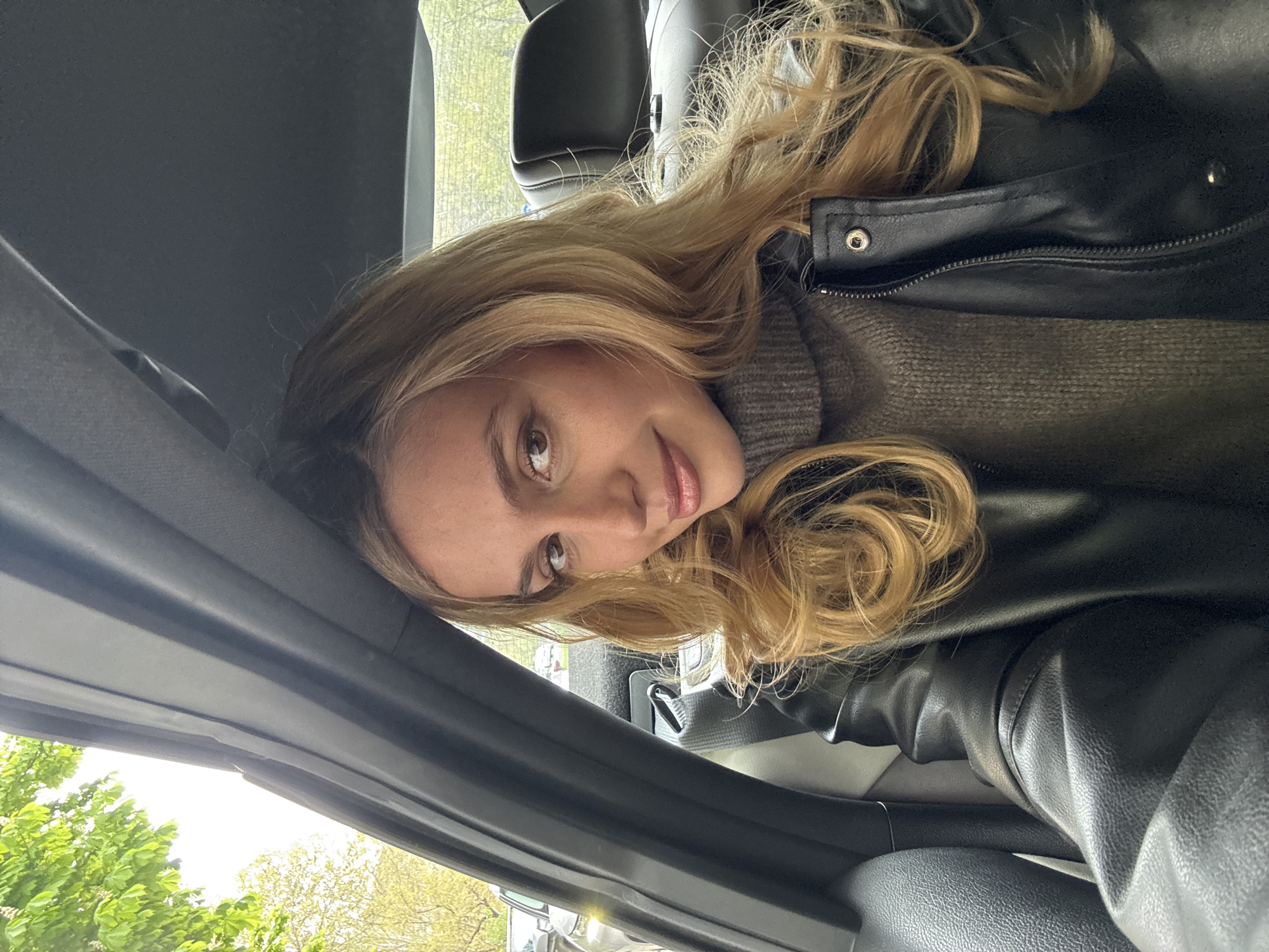 A casual portrait features a young woman inside a car, with the image rotated 90 degrees counter-clockwise. The woman has long, wavy blonde hair and a gentle, soft smile as she looks directly at the camera. She is wearing a grey knitted turtleneck sweater, layered under a black faux leather jacket with silver snap details and a zipper visible on the lapel area. Her head rests against the dark grey interior trim of the car, which includes a car seat, headliner, and door frame elements.
Through the car window, outside elements like lush green trees and a bright, clear sky are visible, suggesting it is daytime with good natural lighting. The light source appears to be from outside, illuminating her face and hair. The car's interior appears dark grey or black. The background outside the window is somewhat blurred, emphasizing the subject. No visible text is present in the image.
