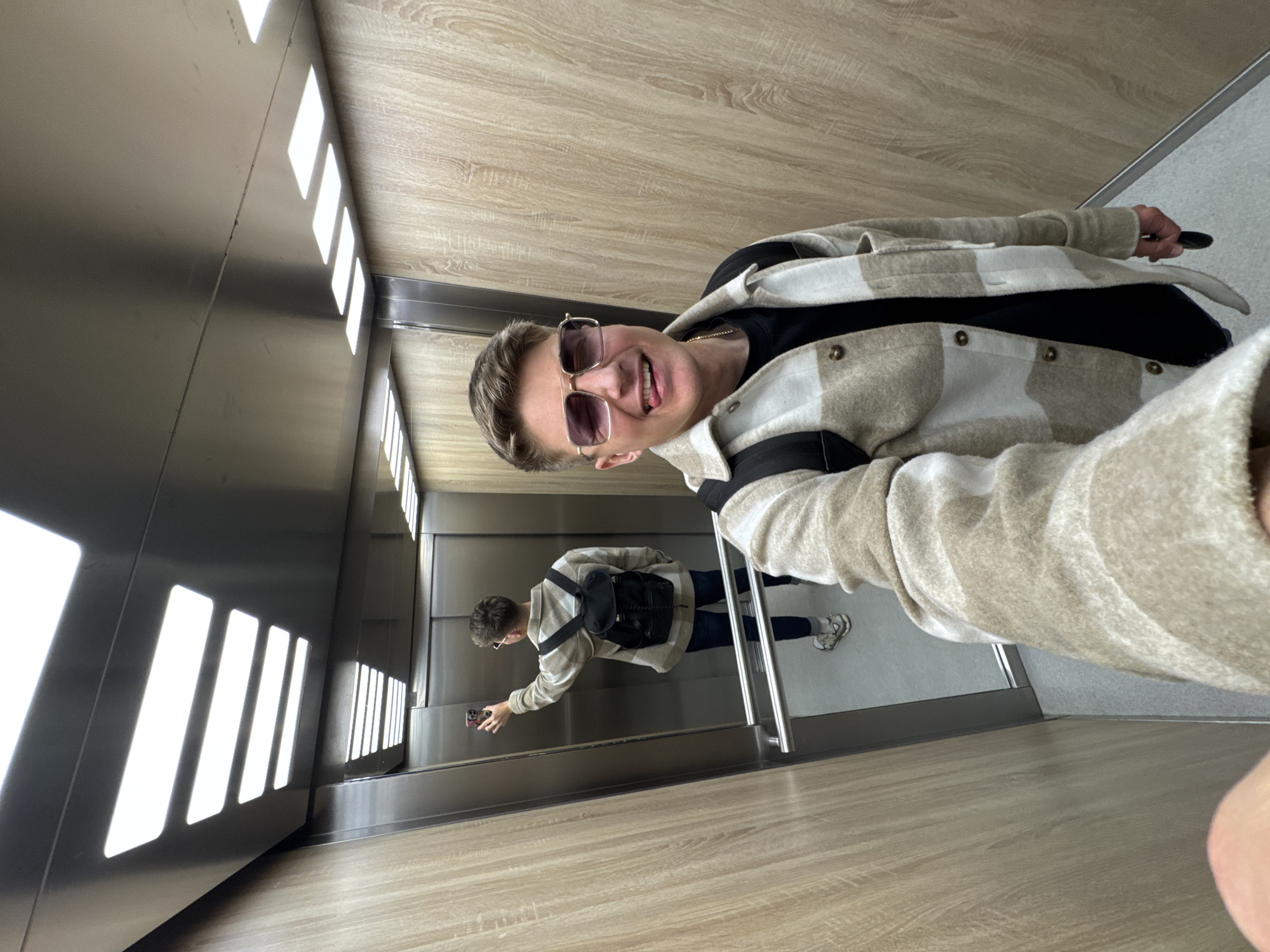 A person is taking a selfie in an elevator. The elevator walls are primarily wood-paneled with a strip of recessed rectangular lights on one side. A mirror on the back wall reflects the person pressing a button. The person in the foreground is wearing sunglasses, a black shirt, and a beige plaid jacket with black straps. The background reflection shows the same person with a black backpack. The location appears to be in a modern building, but without additional context, it is not possible to identify a specific place in Plovdiv, Bulgaria.