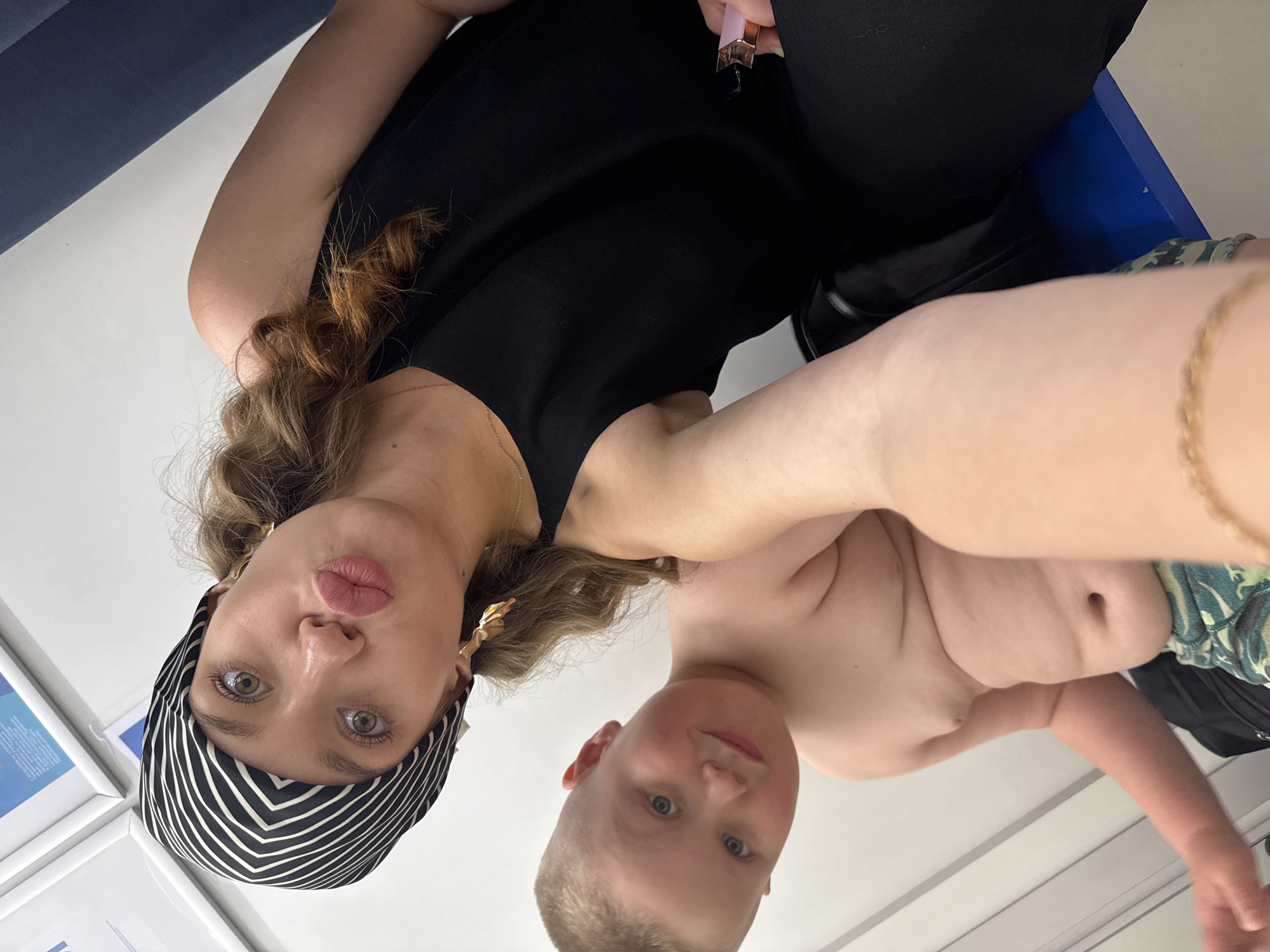 The image shows a selfie taken by a woman with a child. The woman is wearing a black sleeveless top and a black and white striped headscarf. She has on large gold earrings and is making a kissing face. The child, who appears to be young, is shirtless and wearing camouflage shorts. The background appears to be an indoor setting with white walls and a blue chair. There are no discernible landmarks or indications of the city of Votkinsk, Russia.