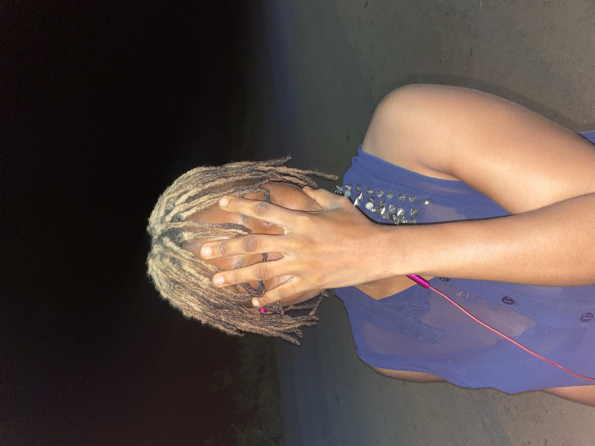 A medium shot depicts a person with dreadlocks obscuring their face with their hands, situated in Ogbe, Nigeria. The person is wearing a sheer, blue, button-down top with studded embellishments. A thin, pink wire, possibly from headphones, is visible draped across their torso. The background is dark, with a gradient of what appears to be a dark road or ground surface. The image is oriented horizontally.