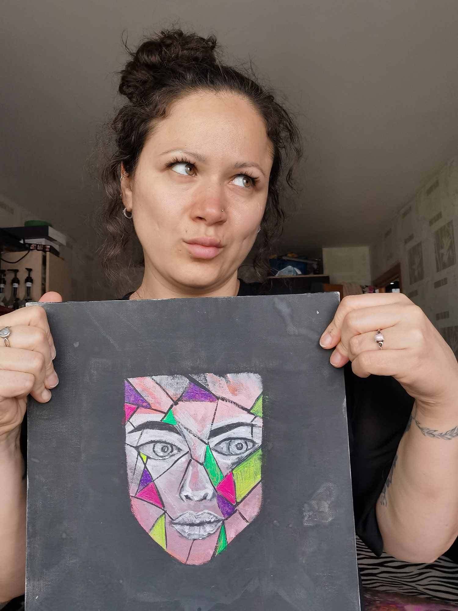 A medium shot captures a woman indoors, holding up a piece of art. She has curly dark brown hair styled in a messy bun and fair skin. Her light brown eyes gaze upwards and to the right, while her lips are slightly pursed in a thoughtful or playful expression. She wears a nose stud and a small silver hoop earring in her left ear. A silver ring with a light-colored stone adorns her left ring finger, and a tattoo resembling a laurel wreath is visible on her left forearm.
The artwork she holds is a square panel, possibly a canvas or board, with a dark grey or black background. On it is a stylized, geometrically segmented face. The face is divided into various shapes colored in shades of pink, purple, green, and yellow, outlined in white or light grey. The eyes are prominent, white with dark pupils, and the lips are also outlined in white.
The background suggests an indoor setting, likely a room in a home in Chaikovsky, Russia. To the left, part of a shelf or cabinet with various items, possibly bottles or containers, is visible. To the right, a wall is covered with patterned wallpaper that appears to have light stripes or a subtle texture. The lighting is soft and diffused, indicating daytime. No visible text is present in the image.