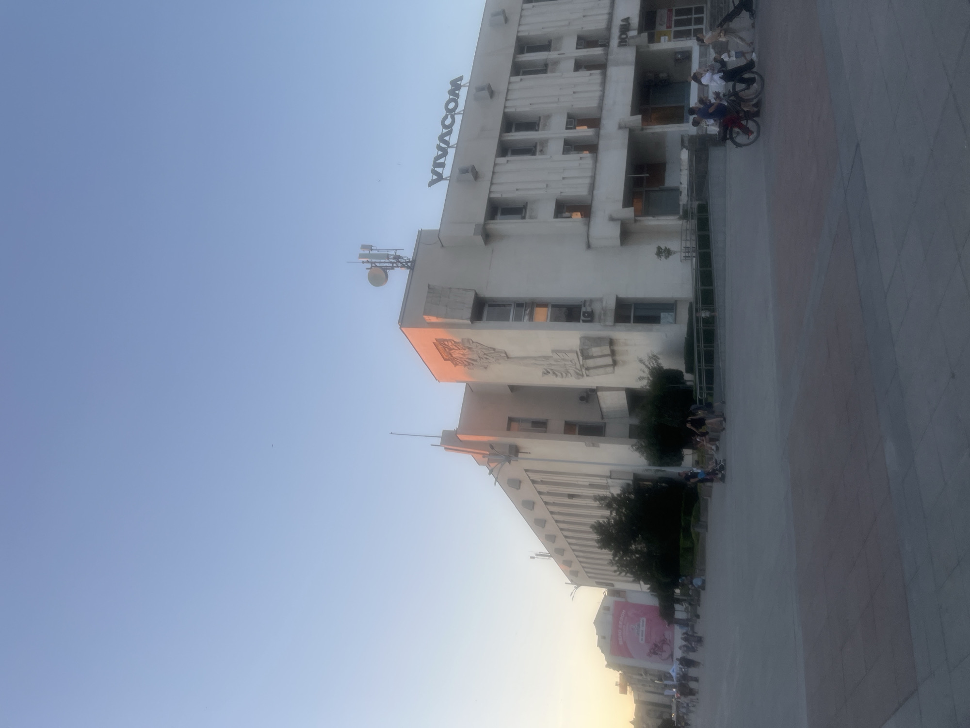 The image depicts a wide, paved plaza in front of a large, multi-story building. The building is light-colored, possibly concrete or stone, and has a modern architectural style with clean lines and geometric ornamentation. The name "VIVACOM" is prominently displayed in large, dark lettering on the upper facade of the building. Above the building, a clear blue sky with subtle hints of sunset orange and pink dominates the upper portion of the image. Several people are scattered throughout the plaza, some walking, others on bicycles or pushing strollers. Trees and bushes are planted in front of the building, and there are streetlights and antennas on the rooftop. A large pink banner with text is visible on a building to the left. The overall impression is that of a public space in a city, likely during late afternoon or early evening.