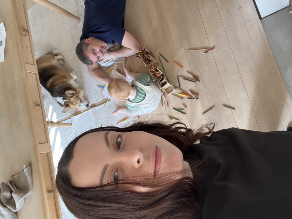 An indoor scene in a domestic residence in Hohenbrunn, Germany, features three individuals and a dog. In the immediate foreground on the left, a woman with long, dark brown hair and a dark-colored top takes a selfie, looking directly at the camera with a neutral expression. Behind her, to the right, a man with short, dark and grey hair, wearing a dark blue t-shirt, lies prone on a light wooden floor, his head propped on his left arm, smiling towards the camera. Next to the man, a young child with reddish-blonde hair, wearing light-colored overalls over a green and white striped shirt, sits on the floor with their back to the camera, appearing to interact with a white sheet of paper and a large quantity of scattered colored pencils. A partially overturned wooden container holding additional colored pencils lies on the floor near the child. Further back, positioned under a light wooden table with visible drawers and knobs, a Pembroke Welsh Corgi dog with tan, white, and dark brown fur lies on its stomach on a white woven rug. The floor is constructed of light-colored wooden planks, and the scene is brightly lit by natural light.