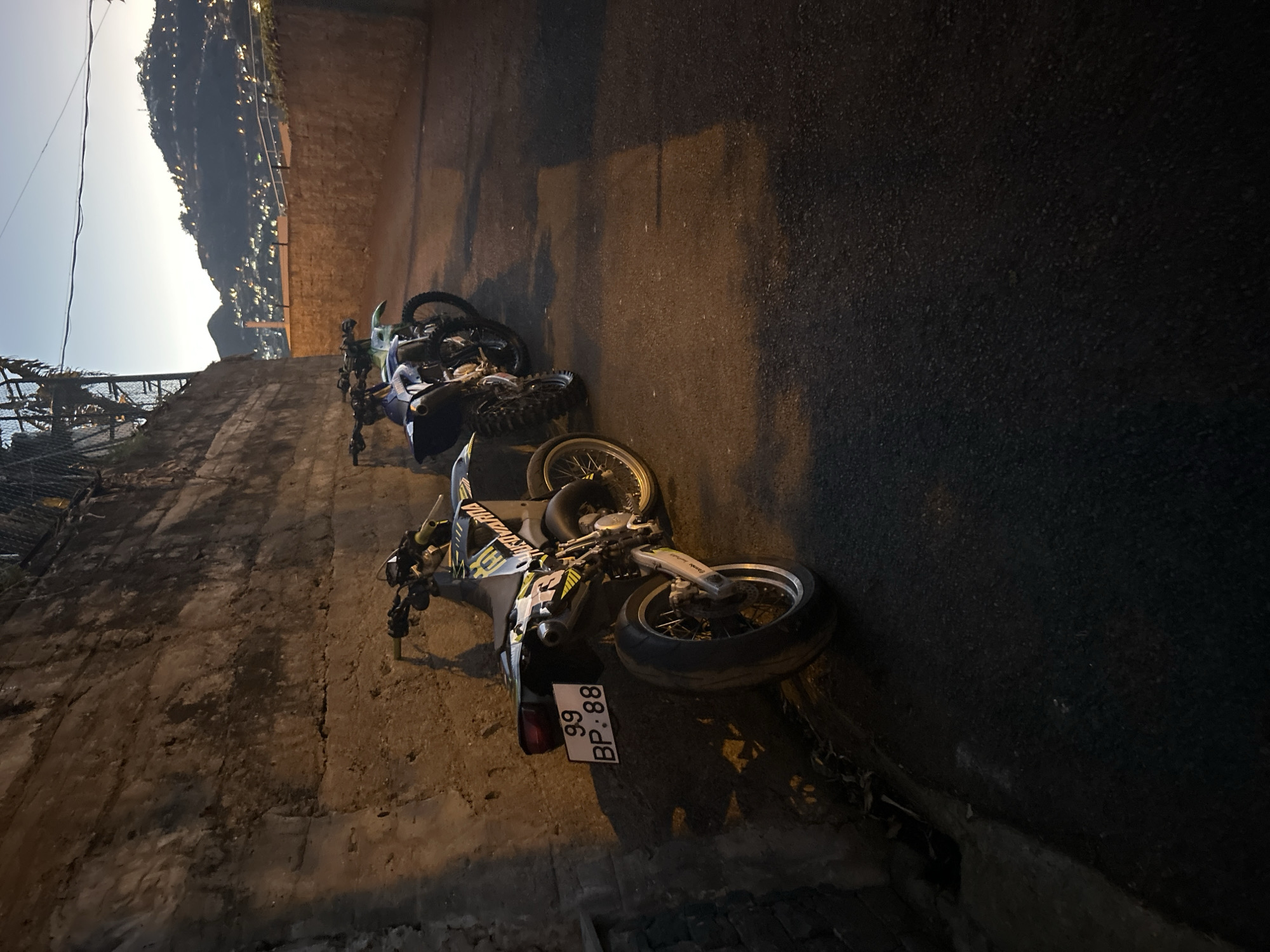 The image shows a street scene in Câmara de Lobos, Portugal. Two motorcycles are parked or lying on their sides next to a wall and a road. The motorcycle closest to the camera is a Husqvarna with a license plate that reads "99 BP 88". In the background is a hillside dotted with lights, suggesting a settlement at night. The lighting in the image is dim and appears to be from streetlights or other ambient sources.