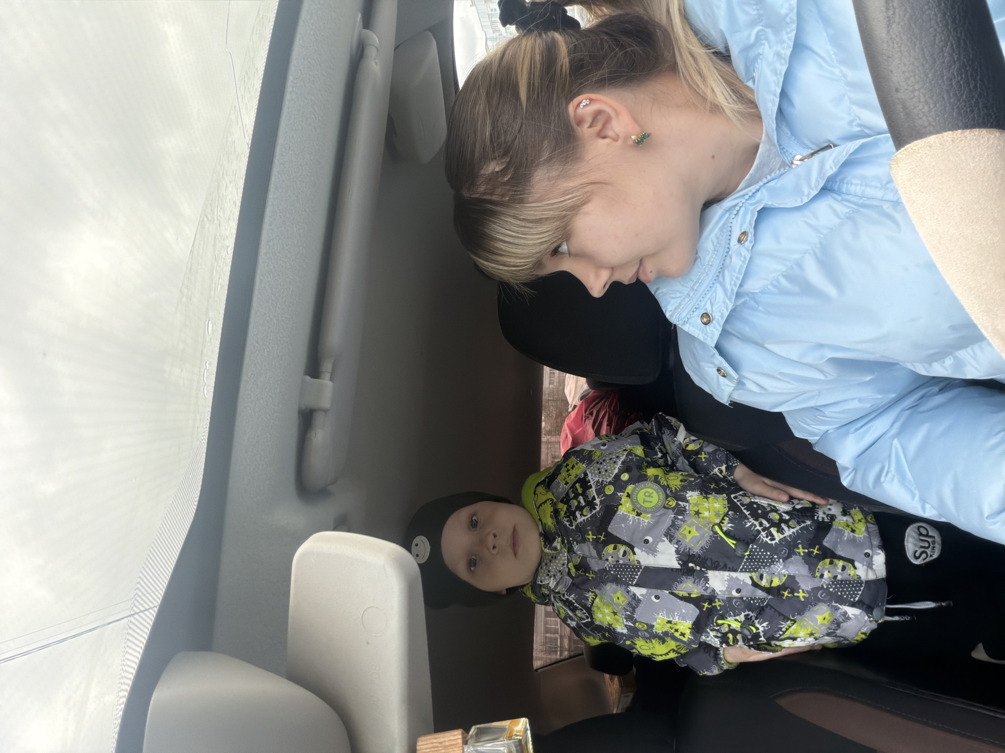 The image shows a woman and a child inside a car. The woman is fair-skinned with blonde hair pulled back in a ponytail, wearing a light blue puffer jacket. She has piercings in her ear. The child is wearing a black hat with a smiley face and a grey and green patterned jacket. They appear to be in a car, possibly parked. Based on the location, they are likely in Chaikovsky, Russia. The car's interior is visible, with the windshield, sun visor, and headrest. There is an air freshener on the dashboard.