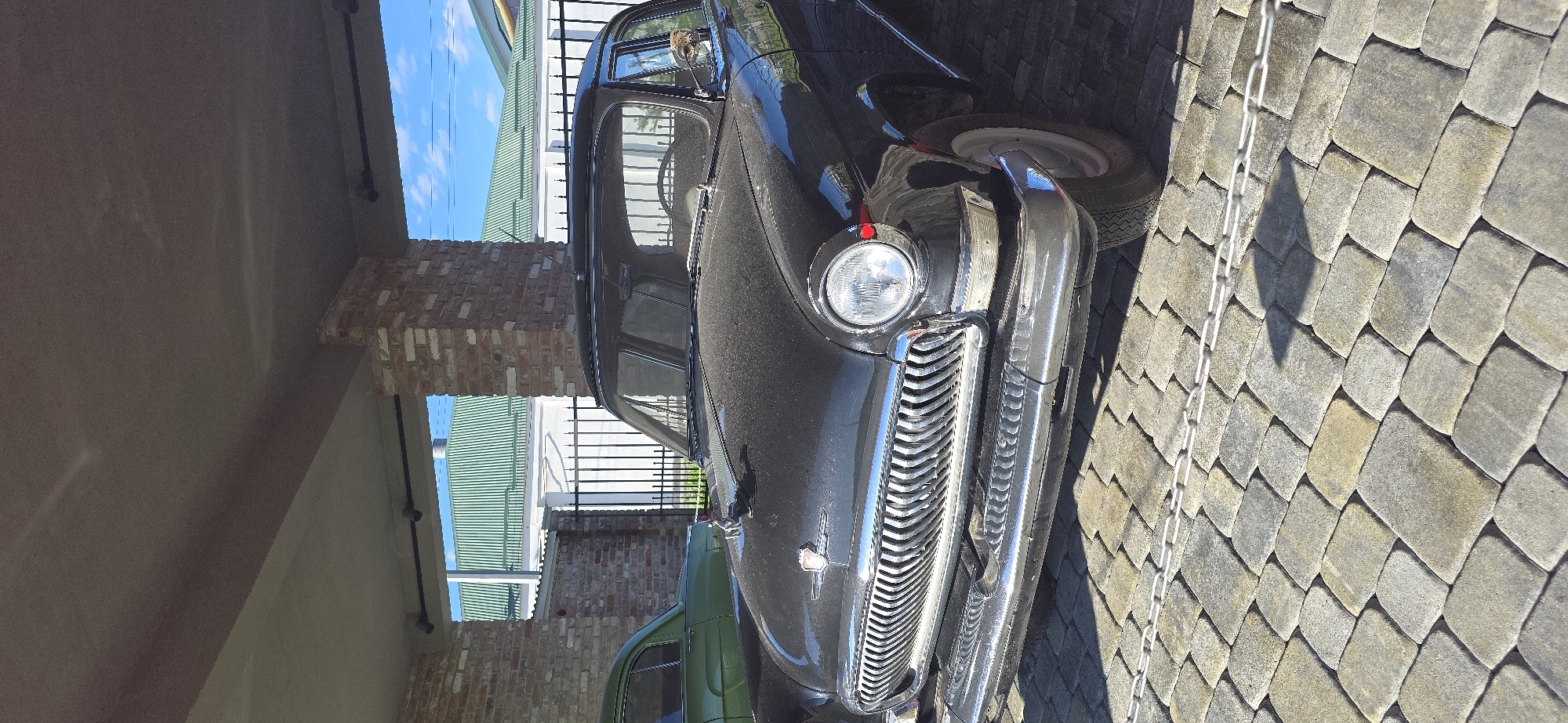 The image shows a black vintage car parked next to a stone pavement. The car is parked on the right side of the image. In the background, a brick pillar and part of a green car are visible. There's also a fence. The location is unknown.