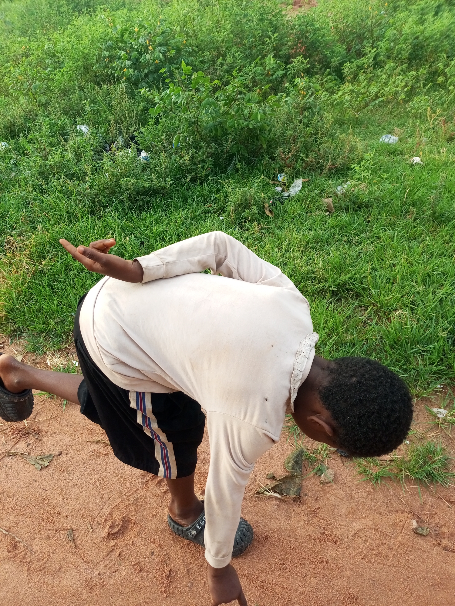 In Tumfure, Nigeria, a young person, likely male based on clothing and hairstyle, is captured in a low crouch on a dirt path. The individual is wearing a light-colored long-sleeved shirt and black shorts with white and red striped trim. Their right leg is extended backward, and their left leg is bent, supporting their weight. The person's head is bowed, with their gaze directed downwards, and their right arm is extended slightly behind them, with the hand held open.
The environment is characterized by a mix of dry, reddish-brown dirt on the path and lush green grass and vegetation in the background. Scattered among the greenery are pieces of white plastic debris, suggesting a degree of littering. The lighting suggests it is daytime, likely late afternoon given the angle of any potential shadows (though not explicitly visible). The overall impression is of an outdoor, possibly rural or semi-urban, setting.