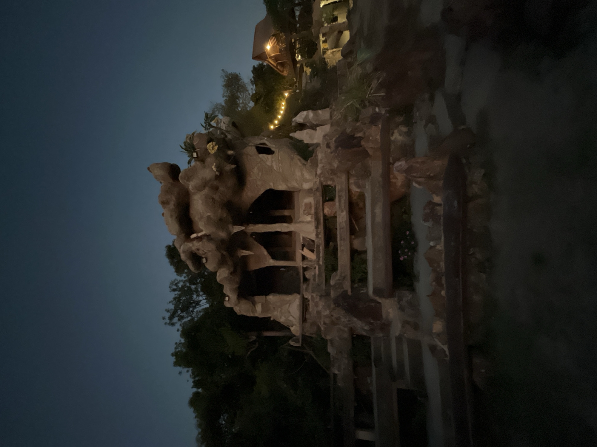 The image depicts a dark, twilight scene in Karimunjawa, Indonesia, featuring a unique, cave-like structure with faux rock formations and a rustic wooden cabin in the background. The cave-like structure, constructed from what appears to be concrete molded to resemble rock, has several openings and is illuminated by subtle lighting, highlighting icicle-like formations hanging from its overhang. A stone staircase with wooden railings leads up to the structure, with some small pink flowers visible in planters. To the right, a traditional Indonesian wooden cabin on stilts is visible, adorned with warm, string lights that create a festive atmosphere. The surrounding environment is lush with trees and foliage, silhouetted against the darkening sky. The overall impression is of a whimsical, possibly themed, establishment set in a tropical location.