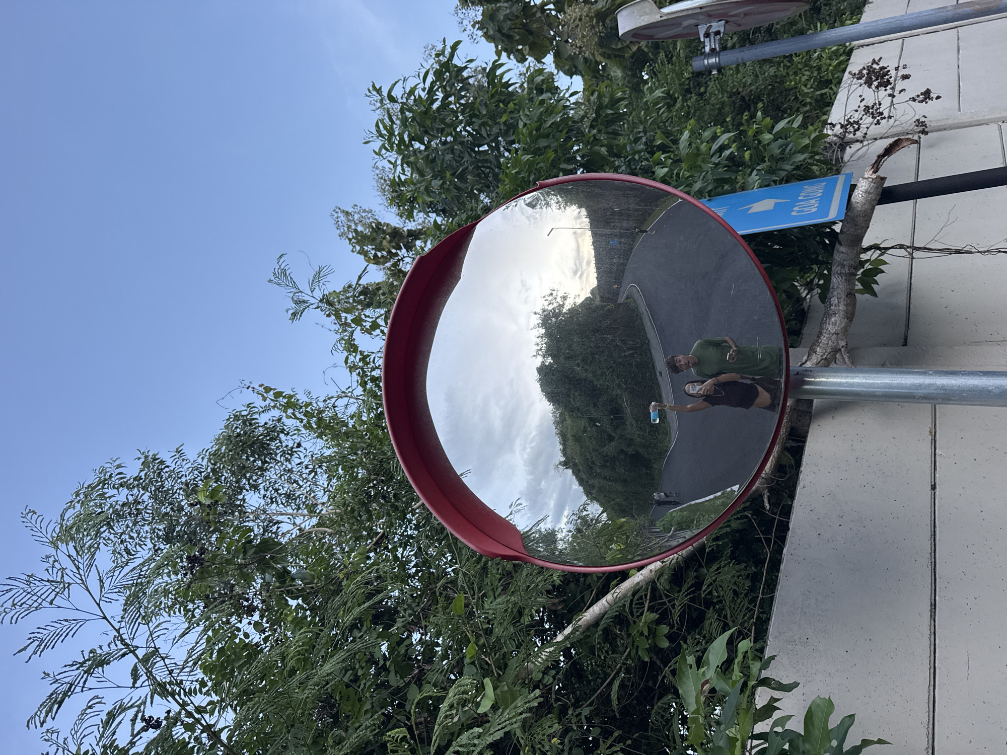 A street scene is captured through a convex mirror. The mirror is red-rimmed and mounted on a silver pole attached to a light grey wall. In the mirror's reflection, a paved road curves away, flanked by dense green trees and vegetation. Two people are reflected in the mirror; one is wearing a green shirt, and another is wearing a brown shirt. One is holding a blue water bottle. A blue sign is attached to the pole above the mirror, pointing towards "GOA GONG". The surrounding environment includes a clear blue sky and lush foliage. The picture appears to be taken in Kuta Selatan, Indonesia, based on the sign directing to Goa Gong.
