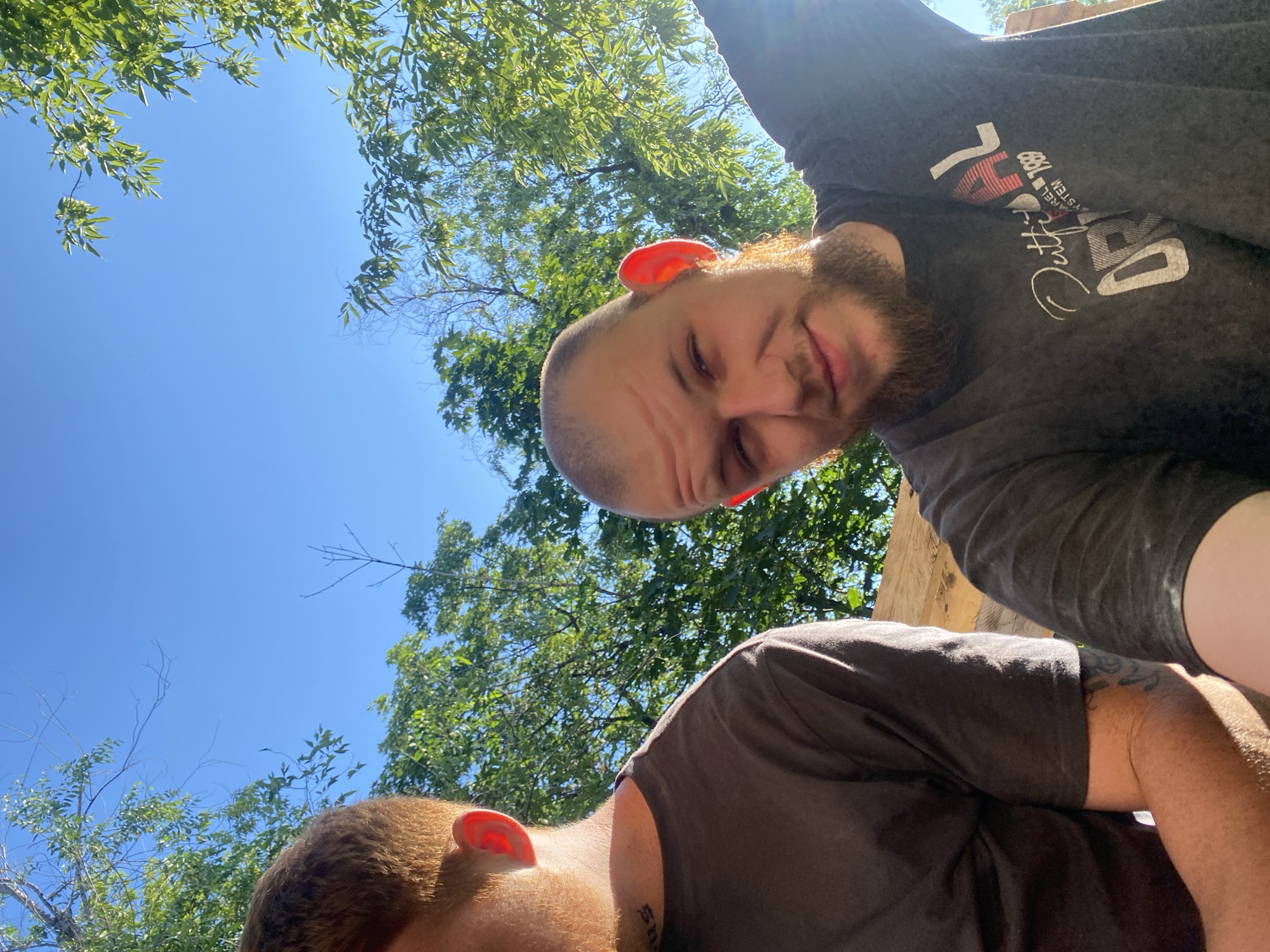 Two men are pictured outdoors, with a clear blue sky and green foliage visible in the background. The man on the right is in the foreground, his face turned towards the camera, with a beard and a shaved head. He is wearing a dark gray t-shirt with text and graphics. His right ear is illuminated by sunlight, appearing bright pink. To his left, the back of another man's head and shoulder are visible. This man has reddish-brown hair and is wearing a dark brown t-shirt. A tattoo is visible on his left forearm. The men appear to be sitting or leaning against a wooden structure. The lighting suggests it is daytime, likely late afternoon due to the warm, directional sunlight. The location is not identifiable as a specific city or region.