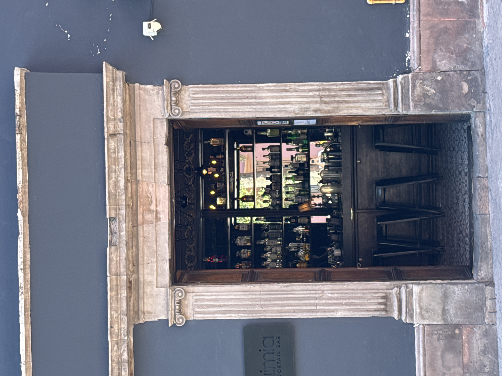 A building with a gray facade features a doorway framed by aged stone and white pilasters with Ionic capitals. Inside, a dark wood cabinet displays an array of glass bottles, possibly a bar. The sign at the bottom left reads "limia COCKTAIL BAR." The scene is located in Querétaro, Mexico.