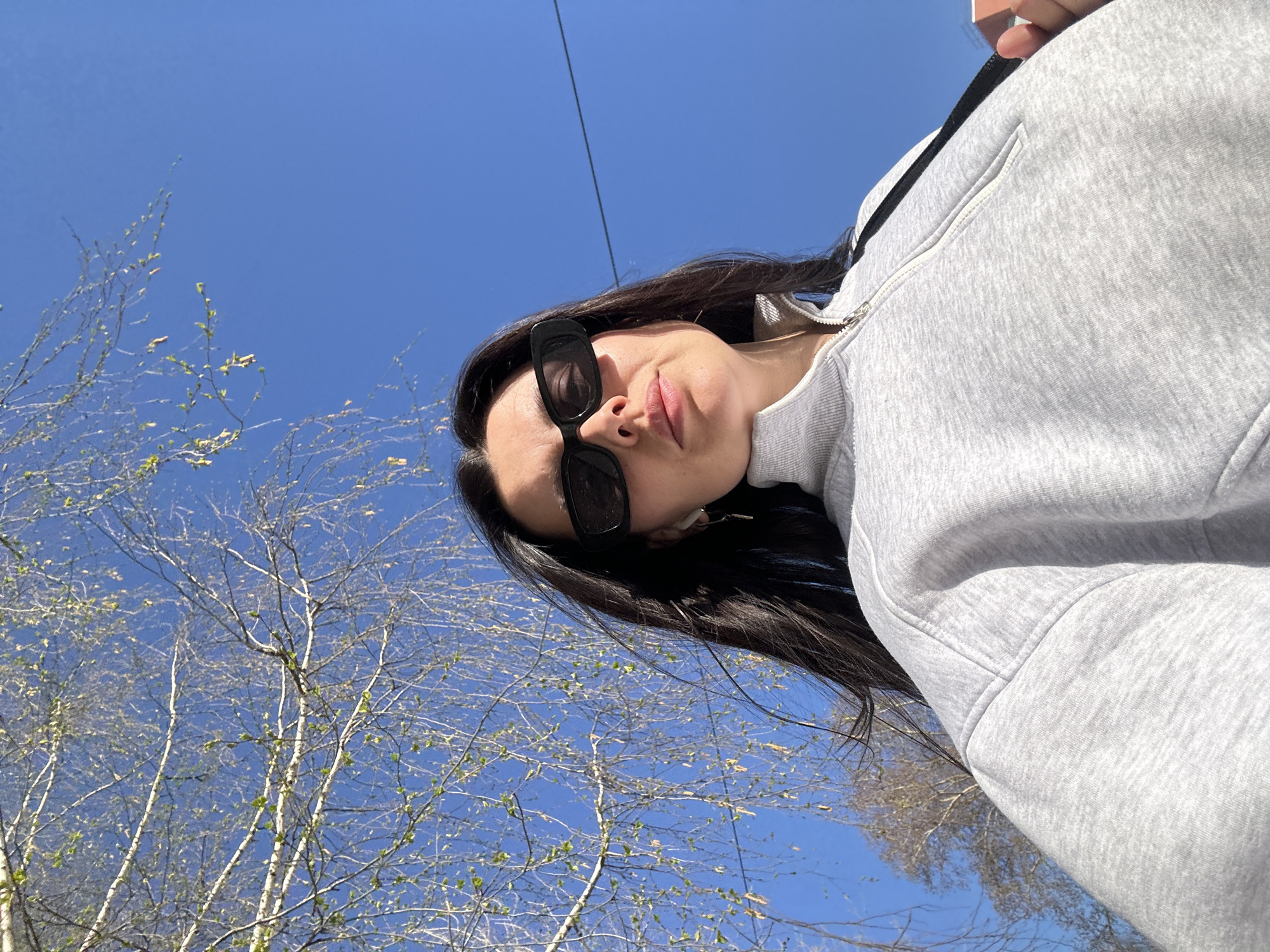A low-angle, outdoor shot shows a woman wearing sunglasses and a light gray zippered sweatshirt, against a backdrop of blue sky and sparse trees, taken in the city of Chaikovsky, Russia. A power line is also visible. The woman is the main subject, with trees and blue sky filling the rest of the frame.