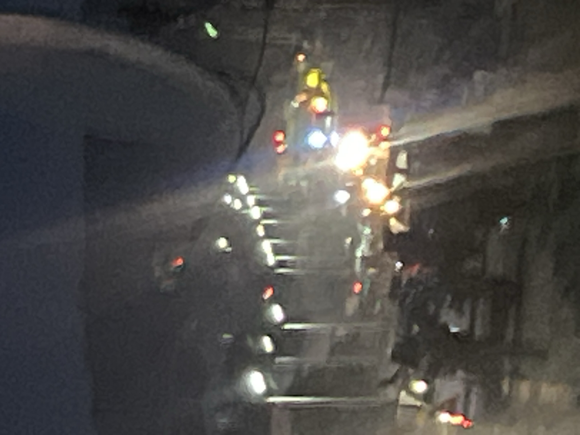 The blurry and dark image depicts an emergency scene at night, illuminated by numerous bright artificial lights. Several figures, presumed to be emergency personnel, are visible amidst the glow. One prominent figure in the upper central portion wears a yellow suit or protective gear, with lights emanating from their person or equipment.
The scene is dominated by various emergency lights, including red, blue, and bright white/yellow sources. A particularly intense and glowing orange and yellow light source is present towards the center-right, suggesting either a fire or very powerful floodlights. A faint haze or smoke appears to emanate from this bright area.
The background consists of indistinct, dark structures. In the mid-left area, a series of horizontal lights suggests a multi-level structure such as a staircase, scaffolding, or part of a large vehicle or industrial facility. The overall impression is one of an ongoing emergency response, characterized by urgency and low visibility. No discernible text is visible within the image. The context indicates the location is Jos, Nigeria.