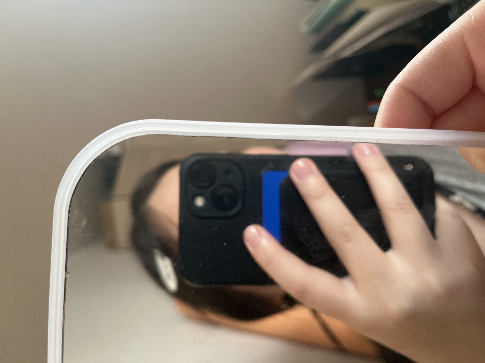 A person's reflection is visible in a rectangular mirror with a white frame, angled to capture a selfie. The reflection shows a person holding a smartphone with a black case, its camera lens visible. The person's hand is in focus, obscuring part of the phone and their face. The background reflected in the mirror is blurred, showing what appears to be an indoor environment with shelves and possibly some objects. The lighting is soft and diffused.
