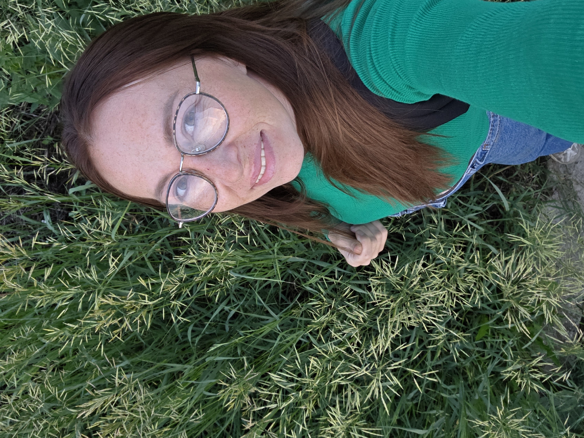 A woman with reddish-brown hair and freckles, wearing round, tortoiseshell-patterned glasses, a green ribbed sweater, and denim overalls, smiles at the camera. She is positioned to the right of the frame, surrounded by lush green grass. The background consists of a dense field of tall grass with lighter green, spiky seed heads. The lighting suggests an outdoor setting, possibly in a park or natural area, during daylight. There are no identifiable landmarks or structures to pinpoint the exact location in Chaikovsky, Russia, beyond it being an outdoor grassy area.