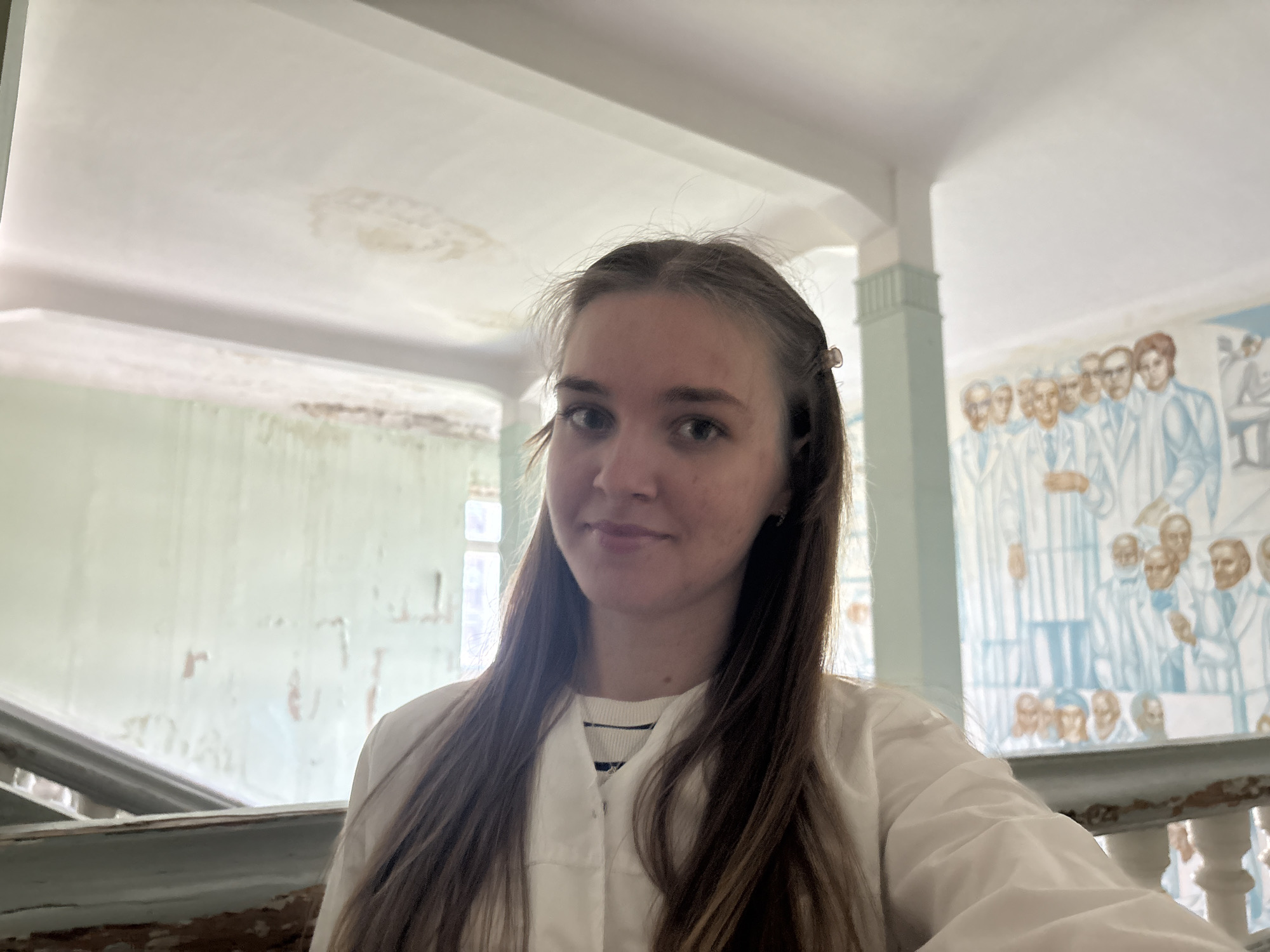 A young, light-skinned woman with long brown hair is the primary subject of this indoor photograph, possibly a selfie due to the angle. She wears a white lab coat over a striped shirt. The background is a dilapidated interior hallway of what appears to be a Soviet-era building in a state of disrepair, with faded light green walls, peeling paint, and stained ceilings. A mural depicting figures in white coats is visible on one wall. The location may be in the city of Perm, Russia.