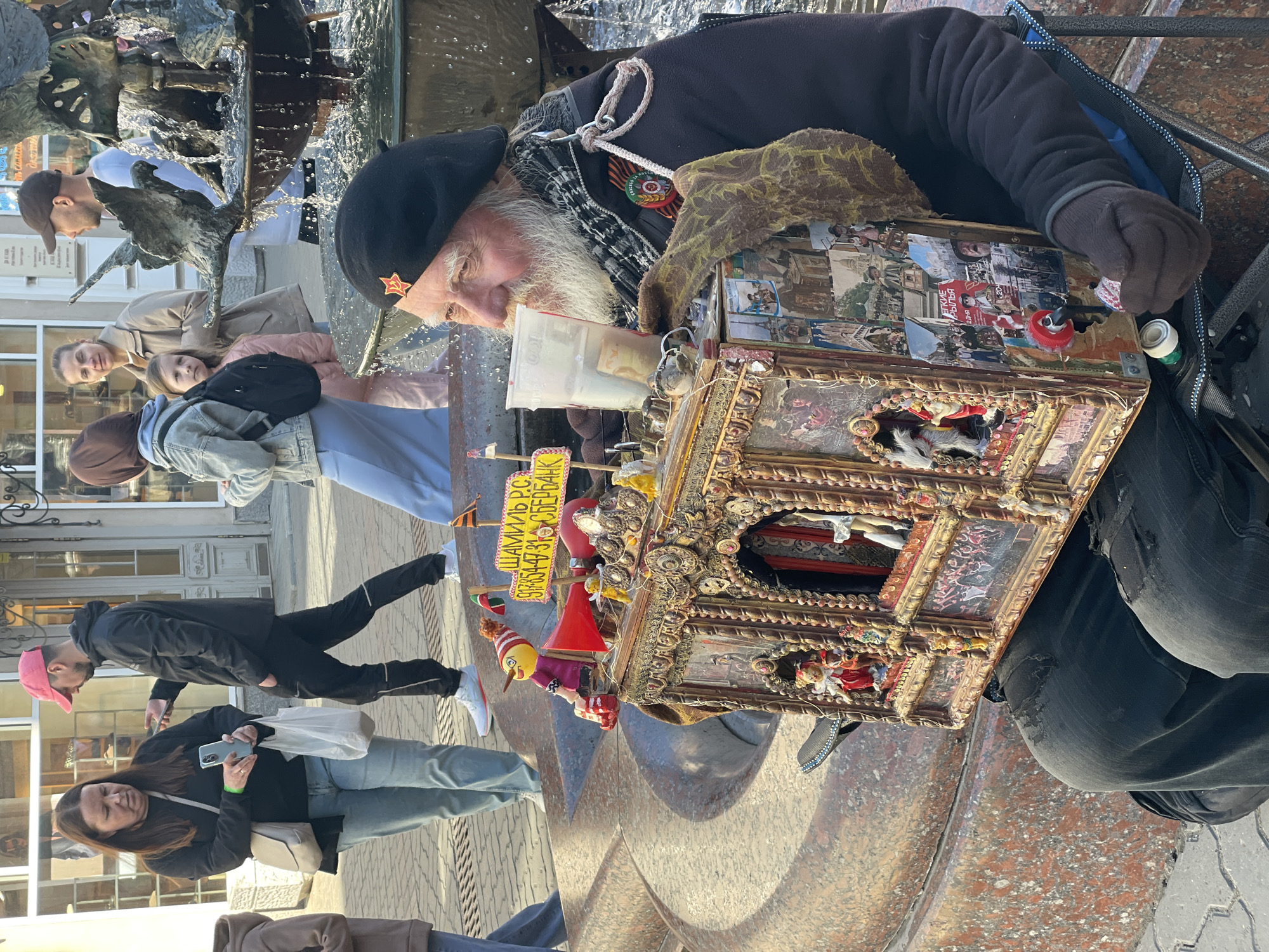 An older man with a white beard, wearing a black beret with a red star, sits with a decorated box on his lap in what appears to be Kazan, Russia. The box is adorned with images, figurines, and small signs, one of which is in Russian. He holds a cup with money in it. He wears dark gloves and a dark jacket. Around him, people walk by and interact with a water fountain. The ground is paved with gray and red stones.