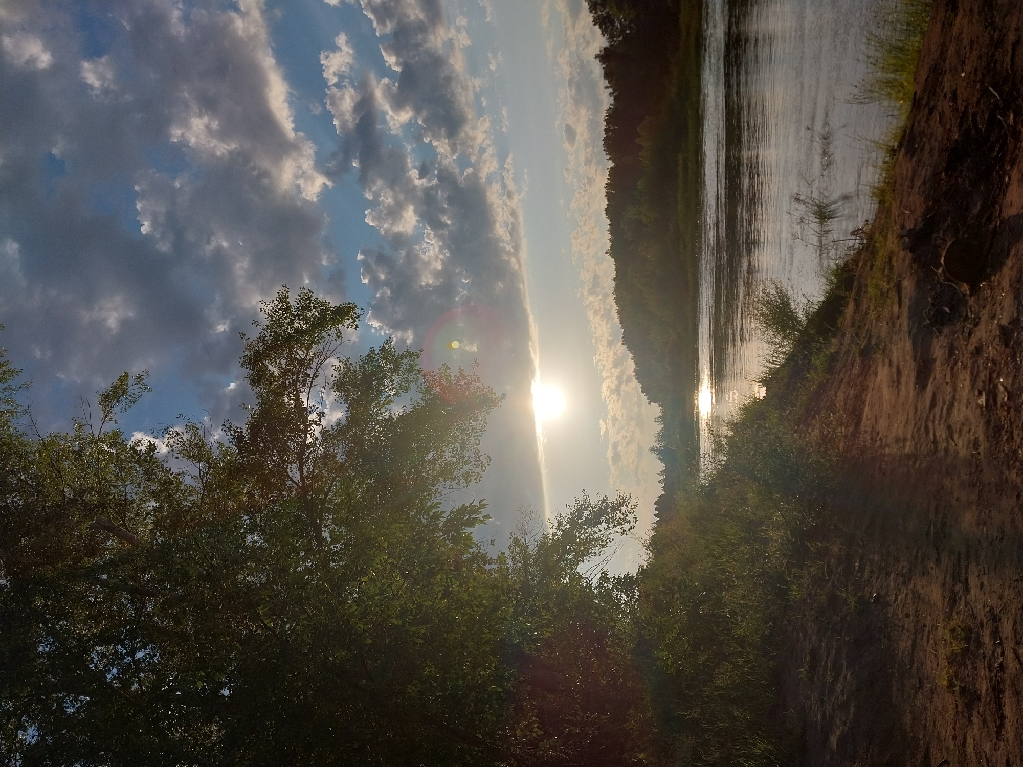 An image captures a serene natural landscape bathed in the warm glow of the setting sun. The scene unfolds along the banks of a tranquil river, its surface reflecting the golden hues of the sky. Lush green foliage drapes over the embankment on the left, with trees reaching towards the heavens. The sky above is a canvas of dramatic clouds, their undersides tinged with grey, contrasting with the soft blue expanse that peeks through. The sun, a radiant orb, is partially obscured by a thin veil of clouds, casting a gentle light that diffuses across the horizon. The foreground is a rough, sandy terrain, hinting at a path leading down to the water's edge. The overall impression is one of peaceful solitude, a moment suspended in time amidst the raw beauty of nature. This scene could be located along the Volga River near the city of Chkalov, Russia, a region known for its picturesque riverine landscapes.