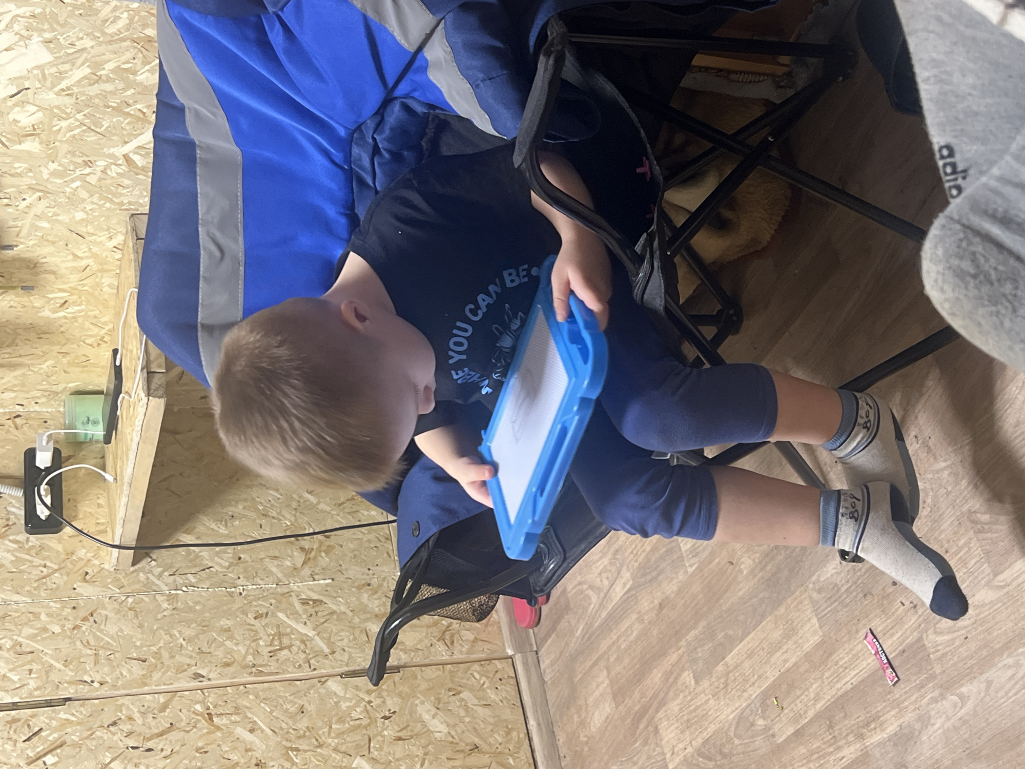 A child sits in a blue and black folding chair, holding a blue-framed drawing board. The child wears a black t-shirt, blue leggings, and socks with blue toes and cuffs. The surrounding environment includes a wall constructed of composite wood panels. An electrical outlet with a charger is affixed to the wall, and wiring is visible. Flooring with a wood-grain design is under the chair. Below the chair, a dog lies on the floor. Based on the composite wood panels and the items on the wall, the location appears to be a room in a residence, possibly in Chaikovsky, Russia.